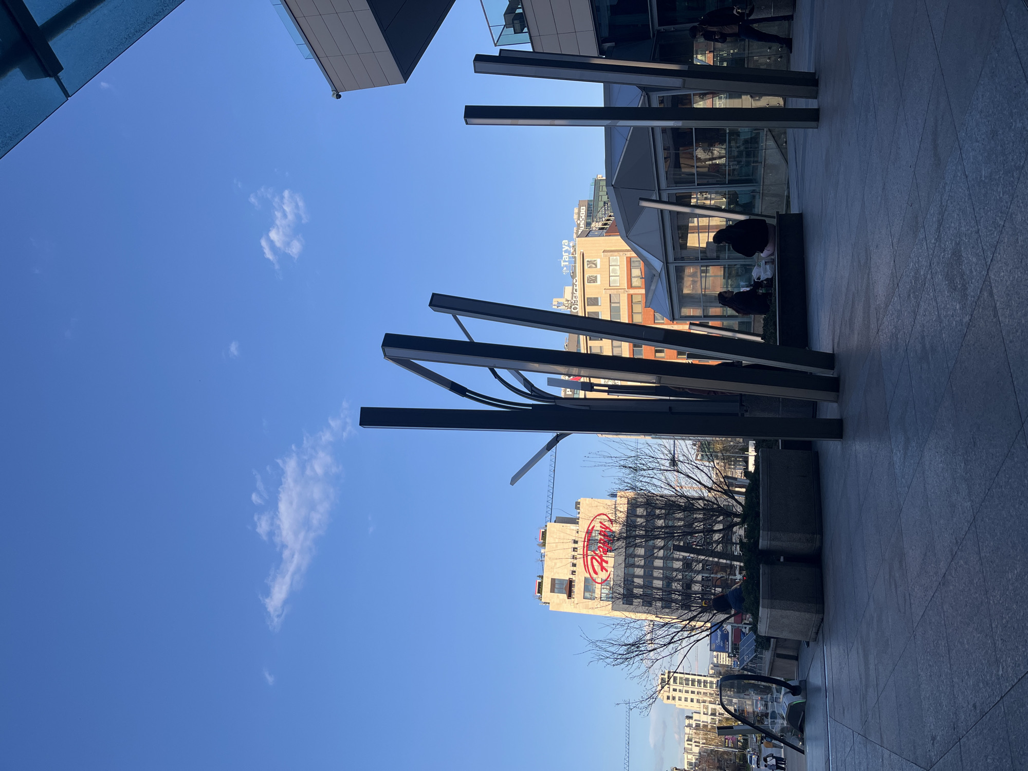 The image depicts a street scene in Sofia, Bulgaria, captured from a ground-level perspective looking upwards. The sky is predominantly blue with scattered white clouds. Modern architecture frames the scene, including sections of buildings with glass and metal facades. A series of linear metal structures are positioned as vertical elements, adding an artistic or architectural detail to the foreground. Several buildings are visible in the mid-ground, one displaying a prominent red logo. Bare tree branches are silhouetted against one of the buildings. People are present, though not prominently featured. The ground is paved with stone or tile.