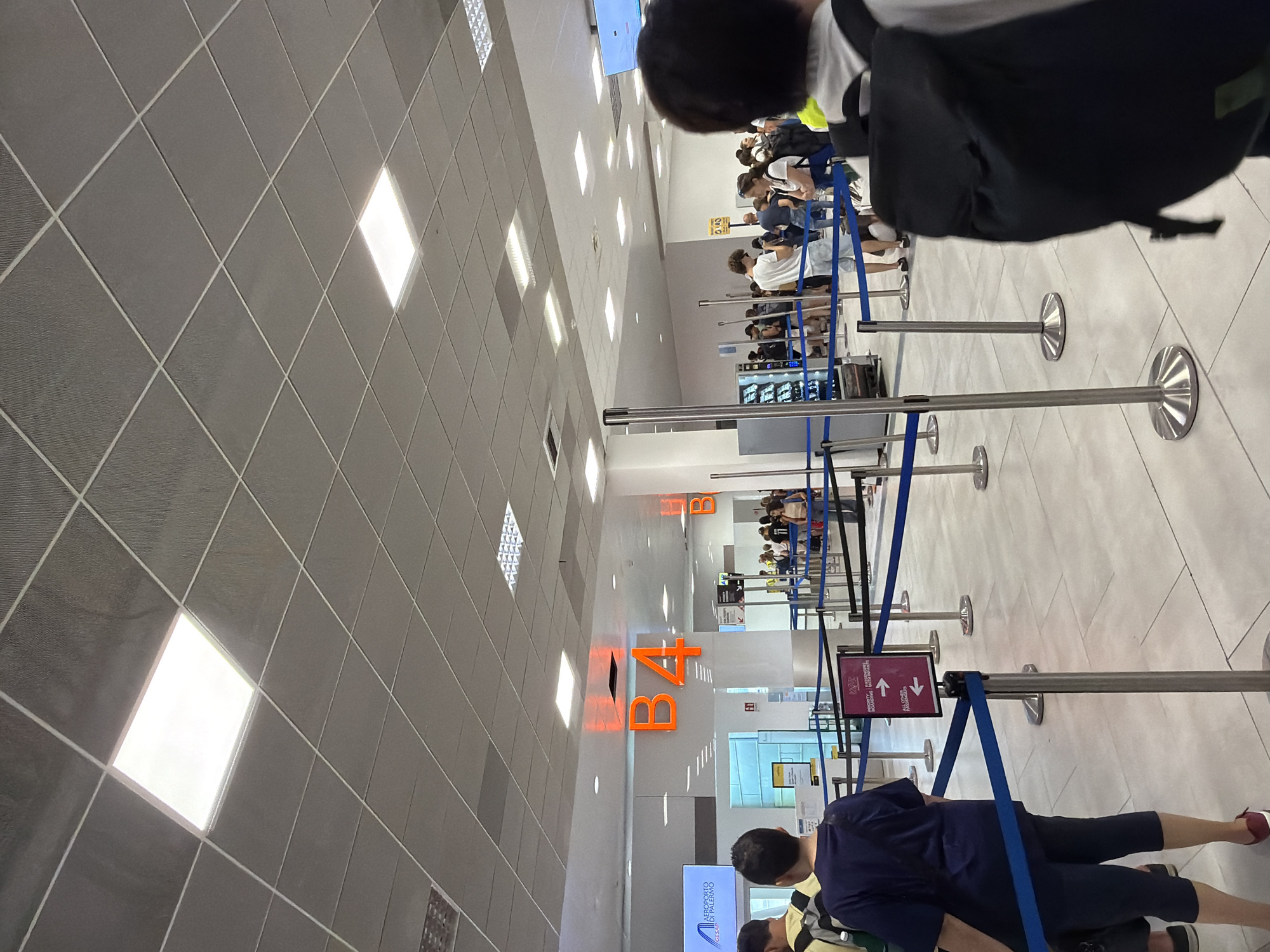 The image captures a scene inside the Palermo Airport in Cinisi, Italy. The airport terminal is brightly lit by a grid of square fluorescent ceiling lights. In the foreground, the back of a person wearing a dark blue t-shirt and dark blue cropped pants, with a black backpack, is visible. They are standing in a queue, with blue stanchion ropes guiding the flow of people. To the left, a sign displays "AEROPORTO DI PALERMO". Prominently displayed overhead in large orange lettering is "B4", indicating a boarding gate or area. Further back, other passengers can be seen queuing near vending machines and at other gates. The floor is tiled with light-colored square tiles. The overall environment suggests a busy airport boarding area during the daytime.