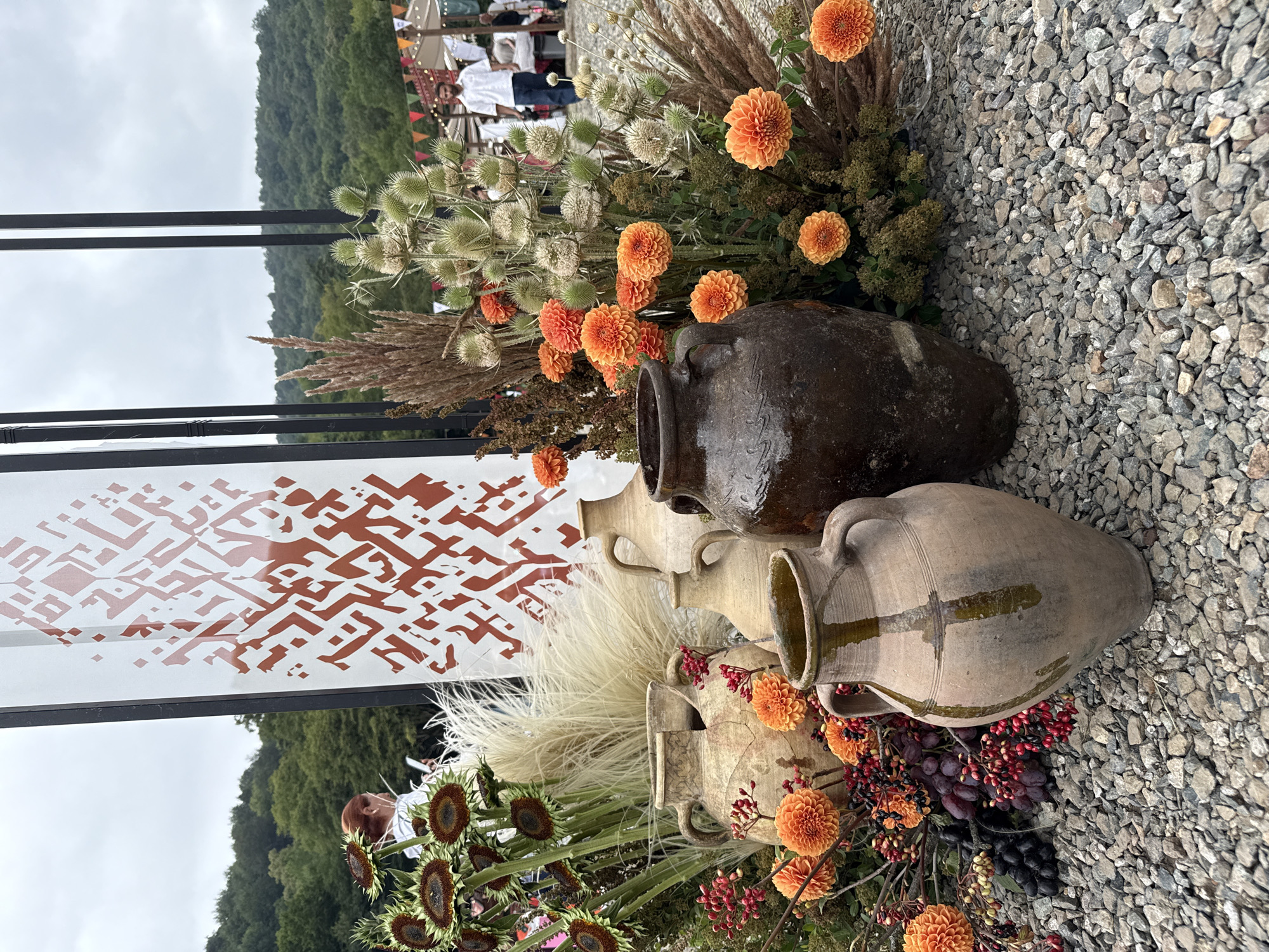 The image depicts an outdoor arrangement of rustic clay pots, flowers, and dried grasses in the city of Dilijan, Armenia. In the foreground, three large, weathered clay pots of varying sizes and colors are prominently displayed on a bed of grey gravel. The largest pot, a dark brown ceramic vessel, sits on the right, while a lighter beige pot with two handles is positioned in the center. A third, taller, beige pot with a greenish glaze is in the front left.
Behind the pots, a vibrant arrangement of orange dahlias, sunflowers with dark centers, and feathery dried grasses creates a naturalistic backdrop. Small clusters of red berries and dark grapes add touches of color and texture. To the left, a woman with red hair is visible in the background, holding a mobile phone. She appears to be looking at the screen.
A tall, white vertical panel with a red, intricate geometric pattern is positioned behind the floral arrangement and pots. The panel is framed by dark vertical poles. In the further background, rolling green hills covered with trees form a scenic backdrop. A man in a white shirt and dark pants walks in the distance, past some tents and stalls, suggesting a market or an outdoor event. The sky is overcast.