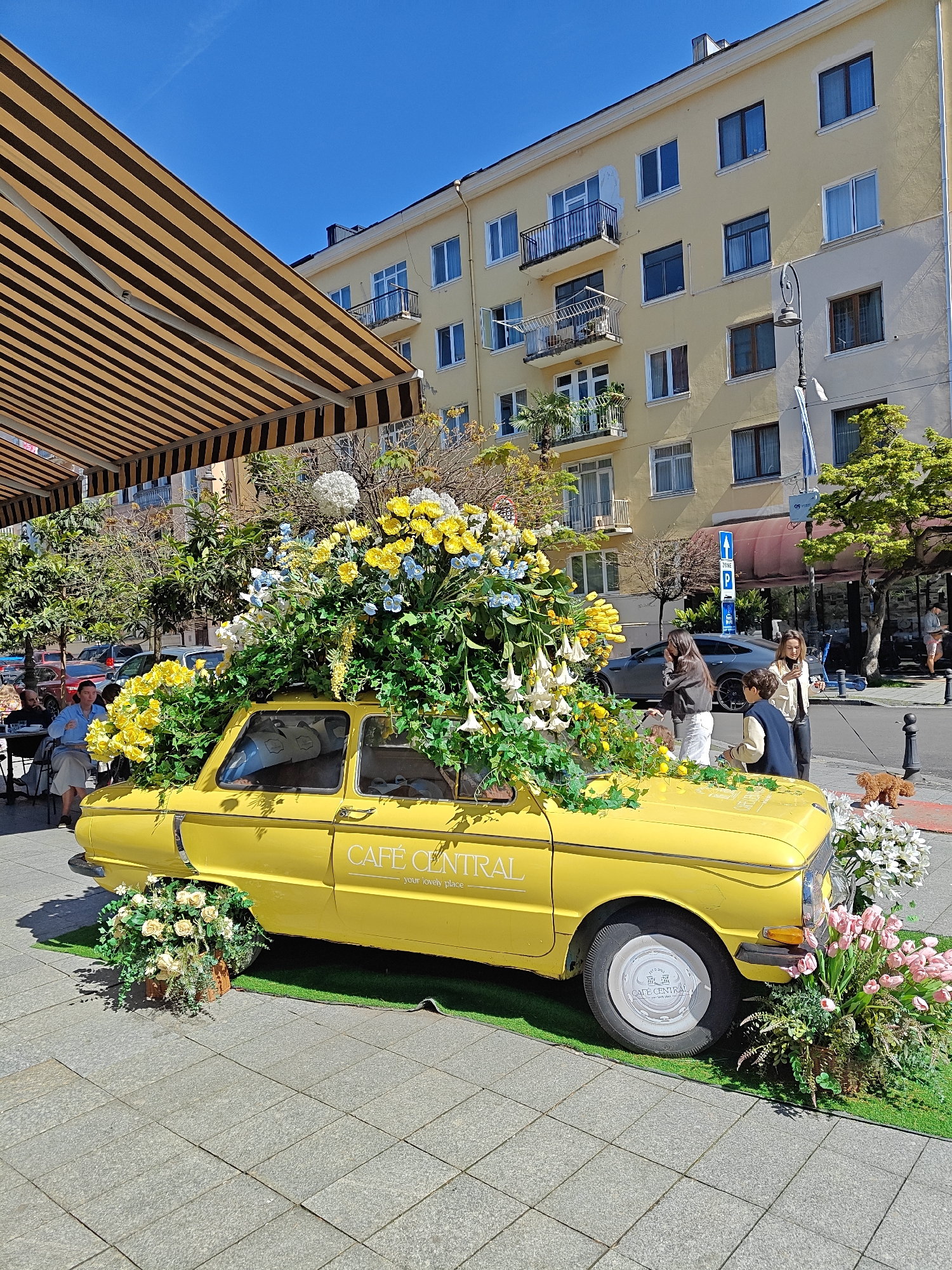 A yellow vintage car, decorated with a profusion of flowers, serves as a Cafe Central advertisement on a sidewalk in Batumi, Georgia. The car is parked on artificial grass, surrounded by flower arrangements. "Cafe Central" is written on the car’s side. Pedestrians are observing the car, including two young women and a child. Tables with patrons are located near the car, under an awning with brown and tan stripes. In the background is a multi-story building, blue sky and trees. A dog is near the right curb.