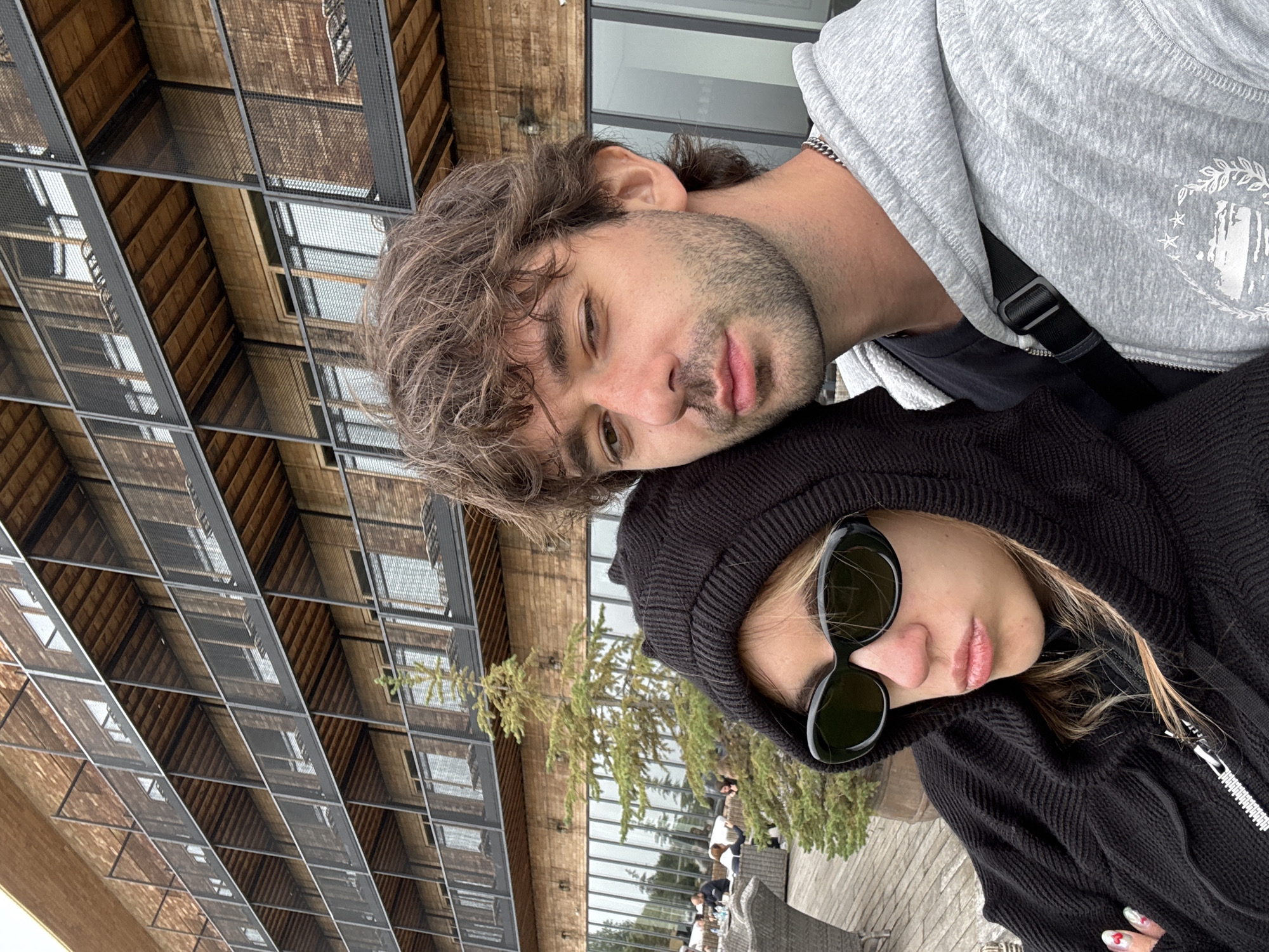 In Stepantsminda, Georgia, a young couple takes a selfie on a wooden deck outside a modern building with a wooden facade and multiple balconies. The woman, on the left, wears a black hooded jacket and dark sunglasses, with her face angled slightly down. The man, on the right, has brown, tousled hair, a light beard, and is wearing a grey hooded jacket over a dark t-shirt. He is looking towards the camera, with his right eye partially closed. A small evergreen tree is situated between the couple and the building. In the background, a few other people are visible seated at outdoor tables. The building features a repeating pattern of wooden slats and balconies with mesh railings.