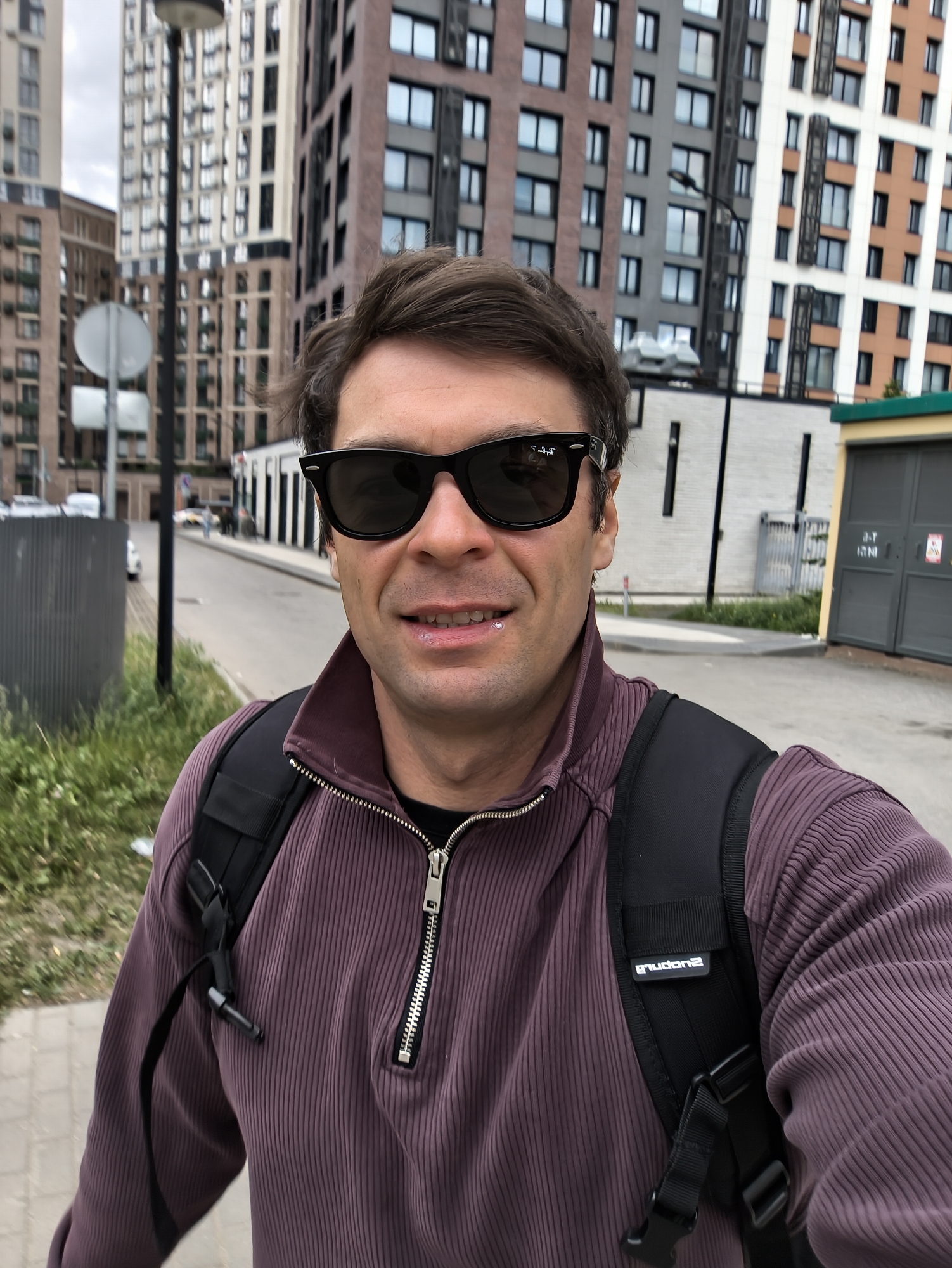 A man stands on the side of a road in a city environment. He is wearing sunglasses, a purple zip-up collared shirt, and a black backpack. Behind him is a street with vehicles, a sidewalk with greenery, and several modern apartment buildings. A street sign is visible on the left. Based on the architecture, the location is likely in Moscow, Russia.