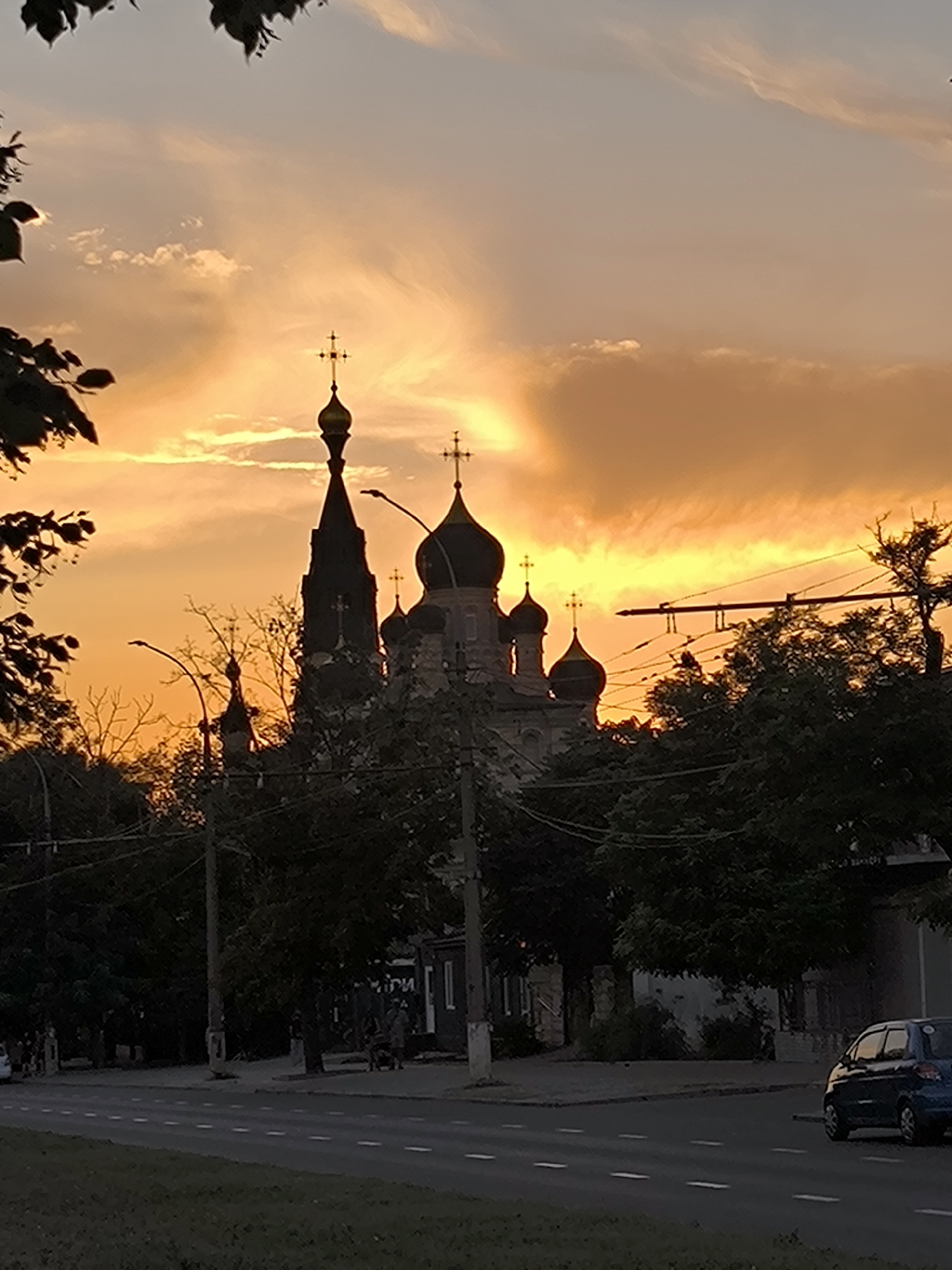 The image depicts a sunset in Mykolaiv, Ukraine, with the sky ablaze in vibrant hues of orange and yellow. Silhouetted against this dramatic backdrop is an Orthodox church, its domes and cross clearly visible. Trees frame the scene, their branches reaching towards the sky, and streetlights dot the landscape. A road runs through the foreground, with a car visible in the lower right corner. The overall impression is one of tranquil beauty and a moment of natural splendor in an urban setting.