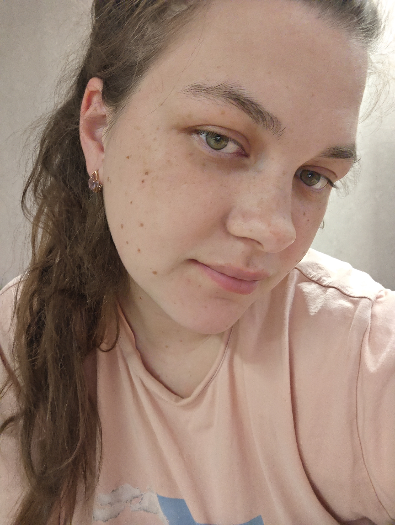 A close-up selfie of a young, light-skinned woman with light brown hair pulled back on one side, exposing an earring in her left ear. She has freckles on her face and neck, and her eyes are green. She is wearing a light pink t-shirt. The background is a plain, light-colored wall. There is no indication of the image being taken in Chaikovsky, Russia.