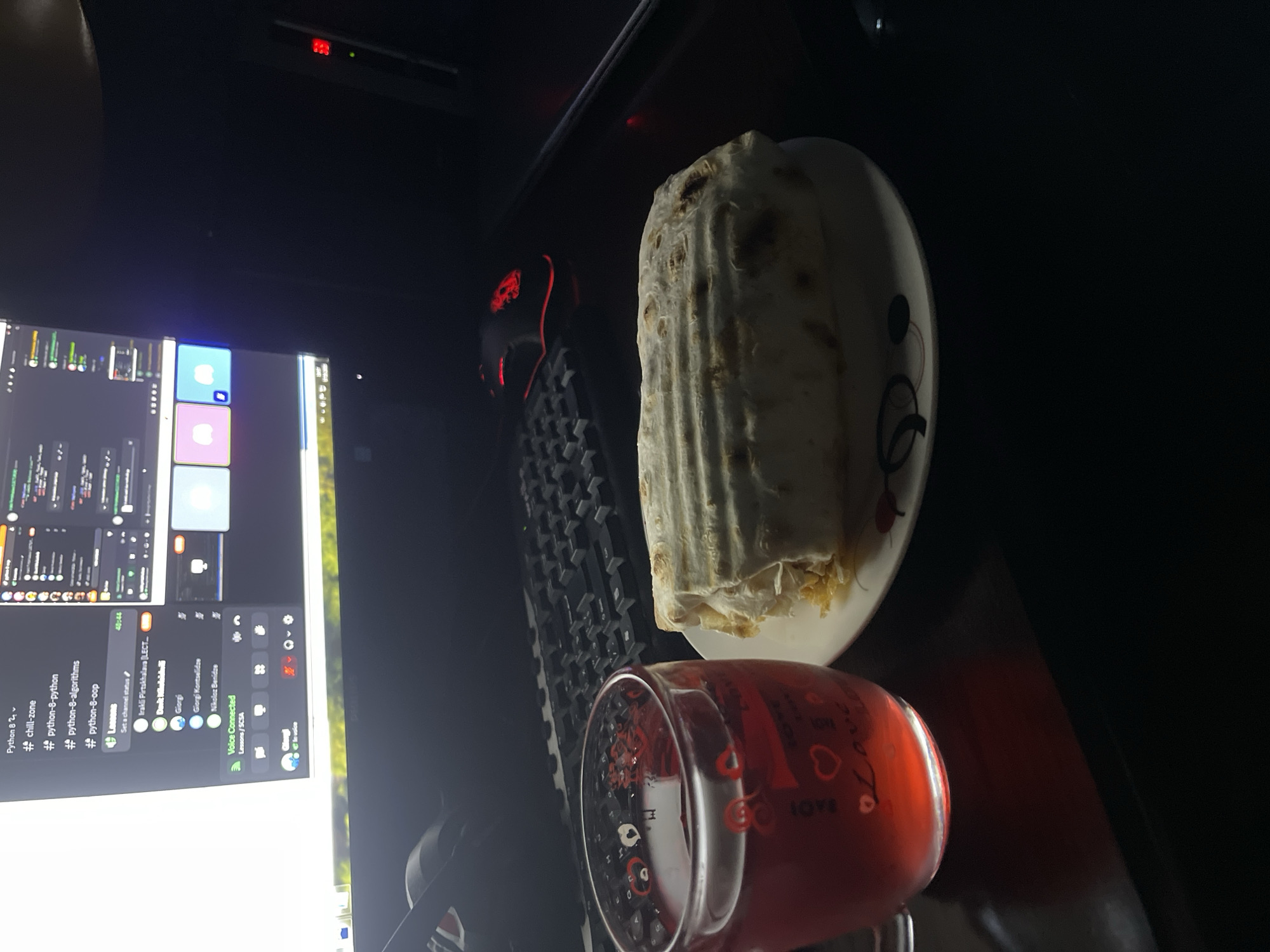 The scene depicts a dimly lit desk setup. A computer monitor on the left displays a Discord window with various programming-related channels, including "Python 8" categories and user mentions. A red gaming mouse and a black keyboard are positioned on the desk. A plate holding a large, possibly wrapped food item sits near the keyboard. A glass filled with a red liquid is located closer to the viewer, adorned with "LOVE" inscriptions and heart designs. The setting appears to be indoors and there are no apparent clues in the image to determine the exact location in Tbilisi, Georgia.