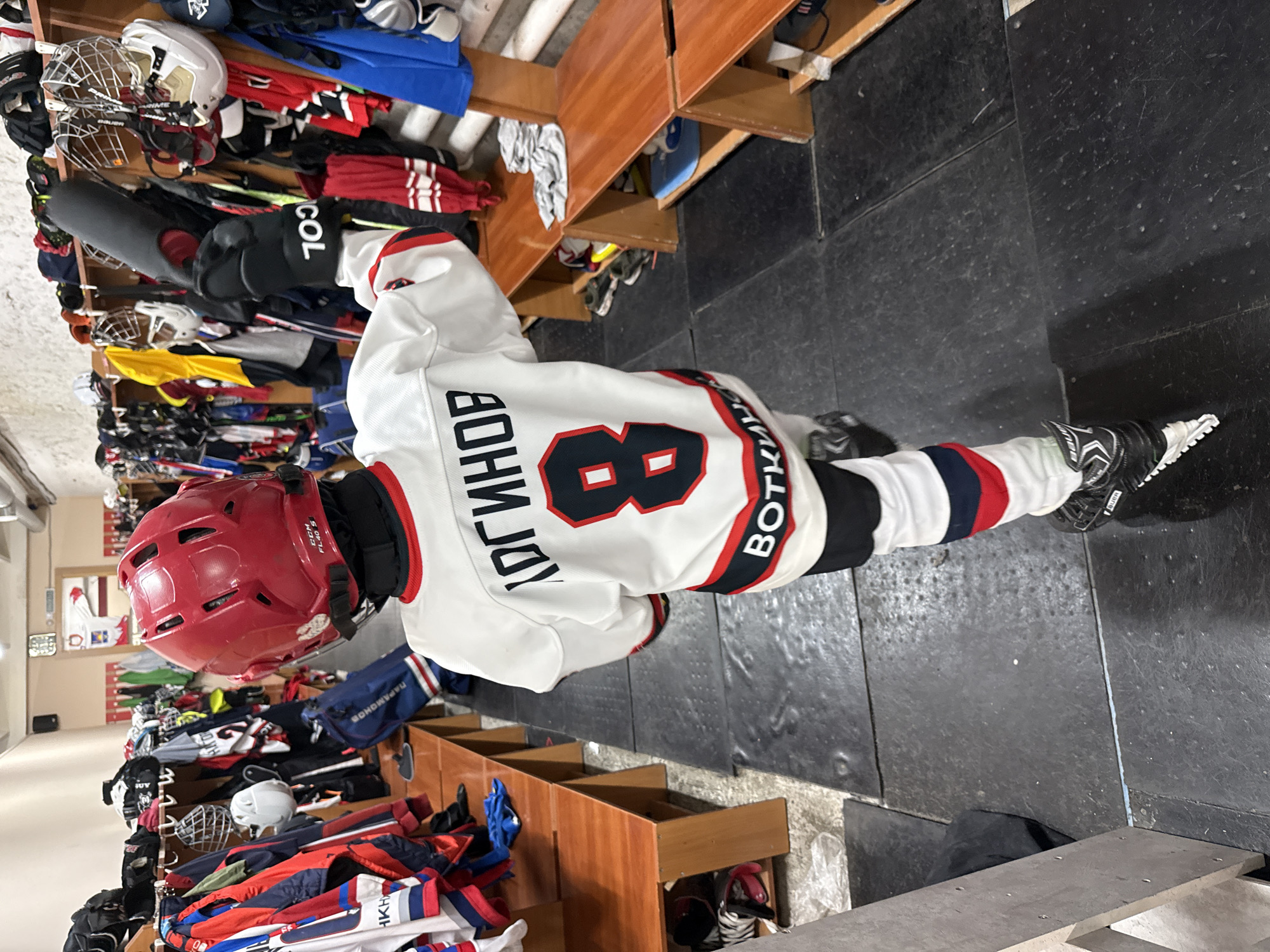 A young ice hockey player, likely a child or teenager, is captured standing in what appears to be a locker room or changing area. The individual is fully dressed in hockey gear, featuring a white jersey with prominent red and black stripes on the sleeves and bottom hem. On the back of the jersey, the name "ЛОГИНОВ" (LOGINOV) is displayed in black letters with a red outline, above a large black number "8" with a red outline. Vertically aligned on the right side of the jersey's back, the text "ВОТКИНС" (VOTKINS) is visible, connecting to the location context of Votkinsk, Russia.
The player wears a red hockey helmet, which has "CCM FL40 S" visible on a sticker. Black hockey gloves are on their hands, with the letters "COL" (likely part of a brand name like CCM) visible on the left glove. Black hockey pants and black ice skates with white blades complete the attire.
The background shows wooden cubbies or benches filled with various pieces of hockey equipment, including multiple helmets, gloves, jerseys, and bags, suggesting a team setting. Brand names like "BAUER" and other text such as "ПАРАМОНОВ" (PARAMONOV) and "AKH" can be partially seen on other gear. The floor is dark, possibly a durable rubber or concrete surface. The lighting is typical of an indoor sports facility. The image is taken from a slightly elevated, side-on perspective, capturing the player standing, ready for or perhaps having just completed a hockey activity.