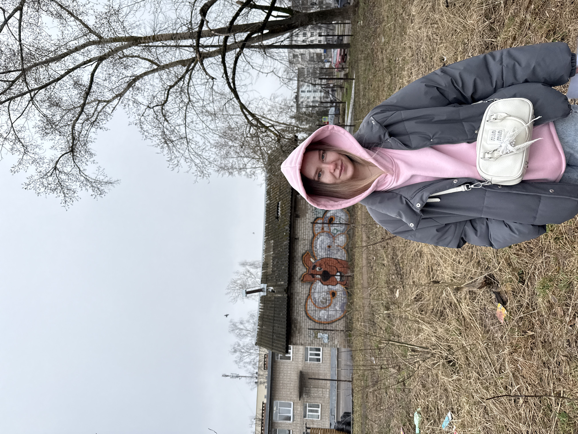 A young woman wearing a pink hooded sweatshirt under a gray puffer jacket and carrying a white purse stands in a grassy field. The background includes a weathered, two-story building with a graffiti mural of a cartoon character on the upper section. The building is gray with white-framed windows. The immediate environment is characterized by dry grass and scattered debris. Bare tree branches are visible above. A multi-story residential building is visible in the distance, indicating an urban or suburban setting. The scene is overcast, with a gray sky. Without further information, the location of this image is unverifiable, including whether it is in Chaikovsky, Russia.