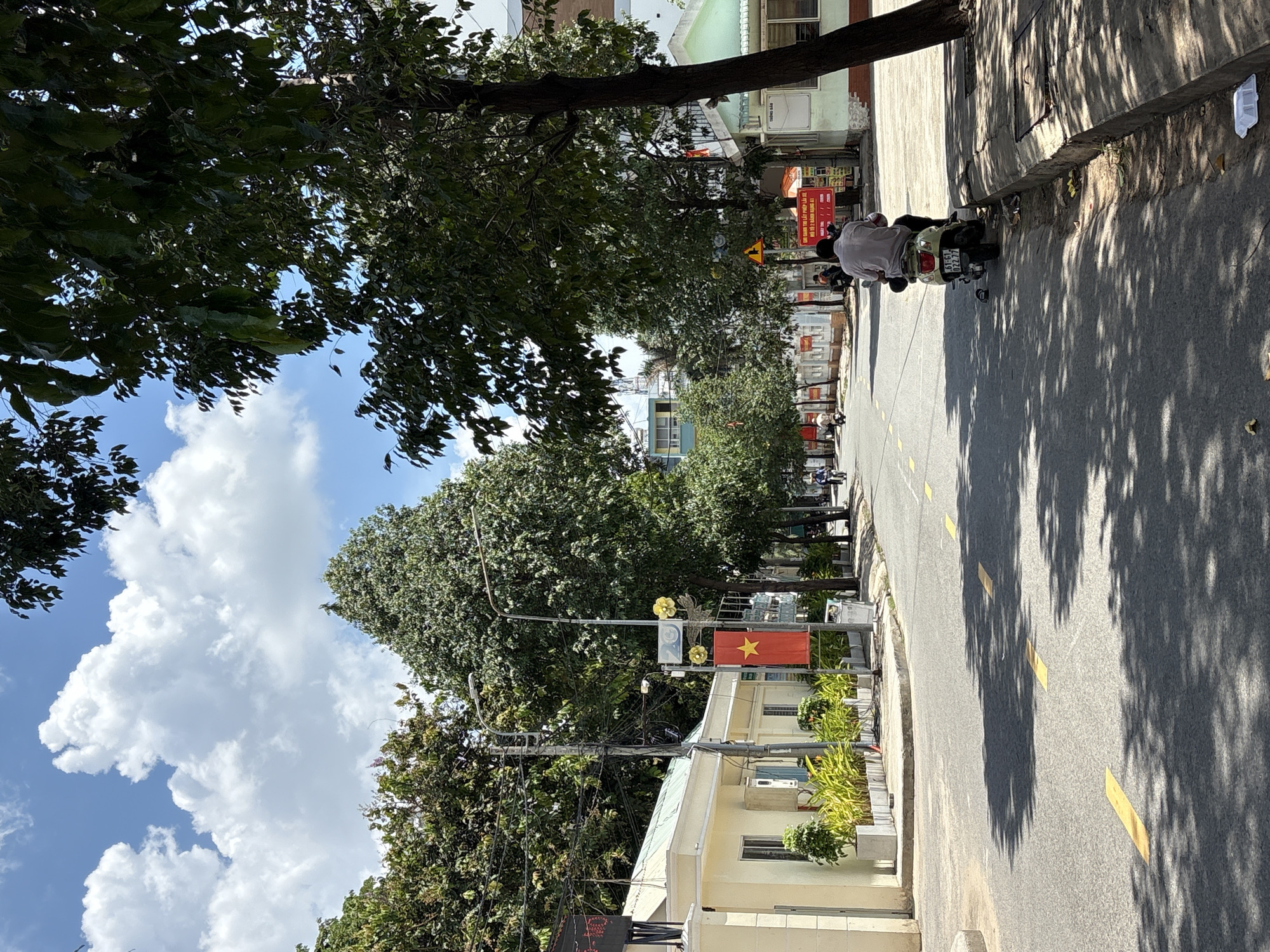 The image shows a street scene in Thuận An, Vietnam. A Vietnamese flag is visible, along with trees that cast shadows on the road. There is a person on a scooter on the sidewalk. The scooter's license plate is partially visible, bearing the numbers "61-61. 022.77". A sign partially visible reads "GHI DANH THỊ LẤY PHÉP LÁI XE KHÔ TÔ 2 BÁNH HẠNG A1". Buildings line the street. The sky is partially visible and contains clouds.