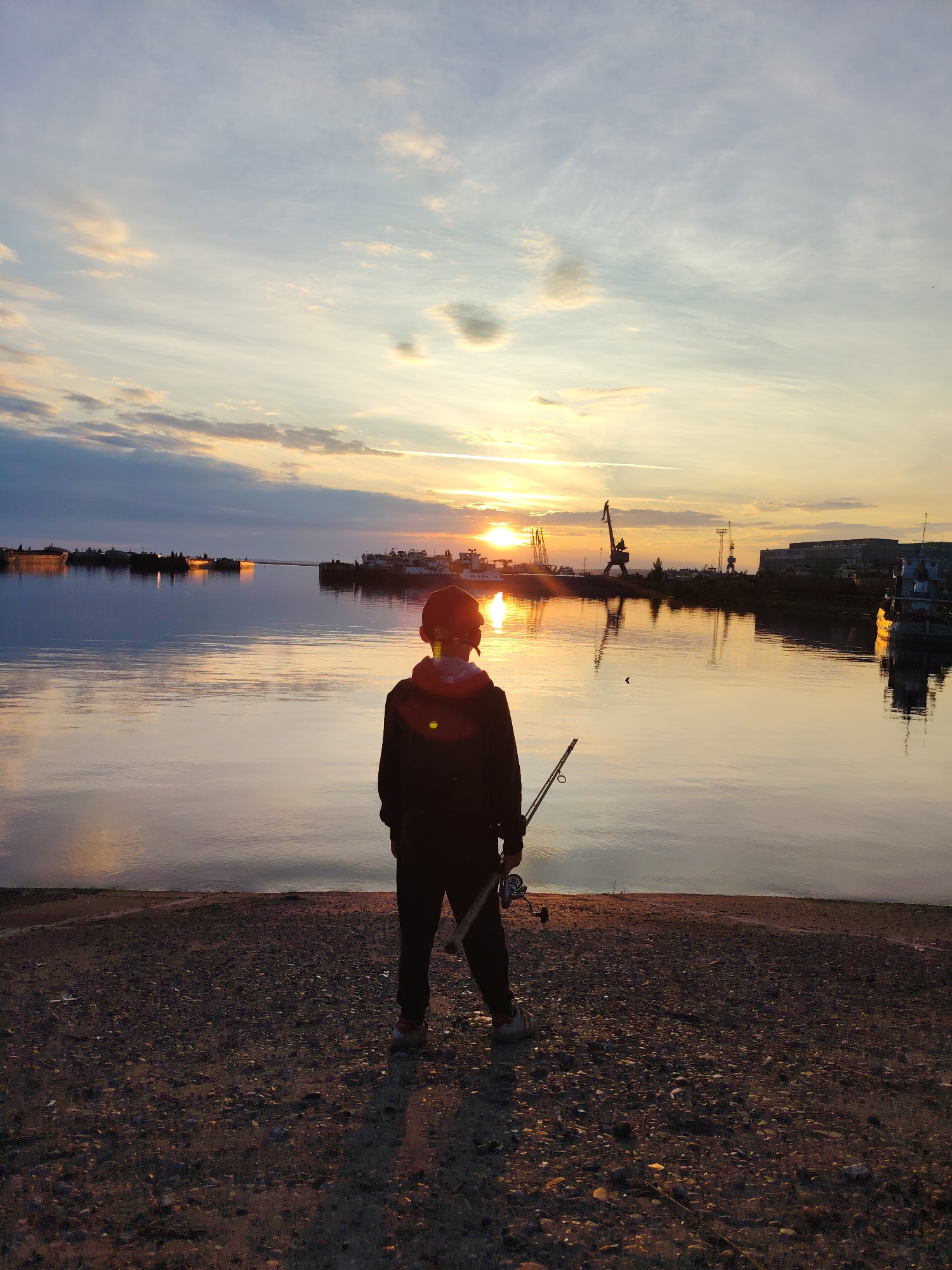 A person is seen from behind, standing on a gravel shore, holding a fishing rod. The person is silhouetted against a sunset sky with scattered clouds. The sun is low on the horizon, casting a warm glow and reflecting on the calm water. In the distance, across the water, a harbor is visible with several docked ships and industrial cranes. This scene is likely located at the Kama River embankment in Chaikovsky, Russia, a city known for its proximity to the river and its industrial port facilities.