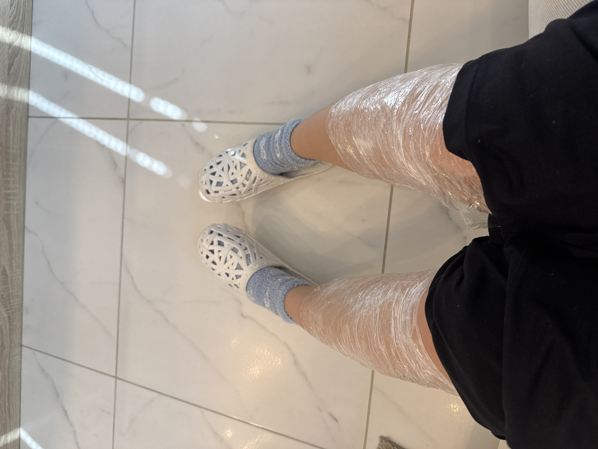 The image displays a first-person, downward view of an individual's lower body within an indoor setting. The subject wears black shorts, light blue socks, and white, latticed slip-on shoes. Both lower legs, extending from just above the knee to the upper ankle, are tightly enveloped in layers of clear plastic cling film, indicative of a body wrapping procedure. The visible skin is light-toned. The flooring consists of large, polished white tiles with subtle grey marbling and light-colored grout lines forming a rectilinear pattern. Two parallel, elongated streaks of bright sunlight are cast upon the floor in the upper half of the frame. A blurred section of light-colored wooden flooring or trim is visible at the top edge, and a small portion of a light-colored, possibly upholstered, surface appears in the bottom right, suggesting the individual is seated. The image does not contain specific visual identifiers to pinpoint a precise location within the City of Zagreb, Croatia; however, the environment is consistent with a private residence, a spa, or a beauty salon.