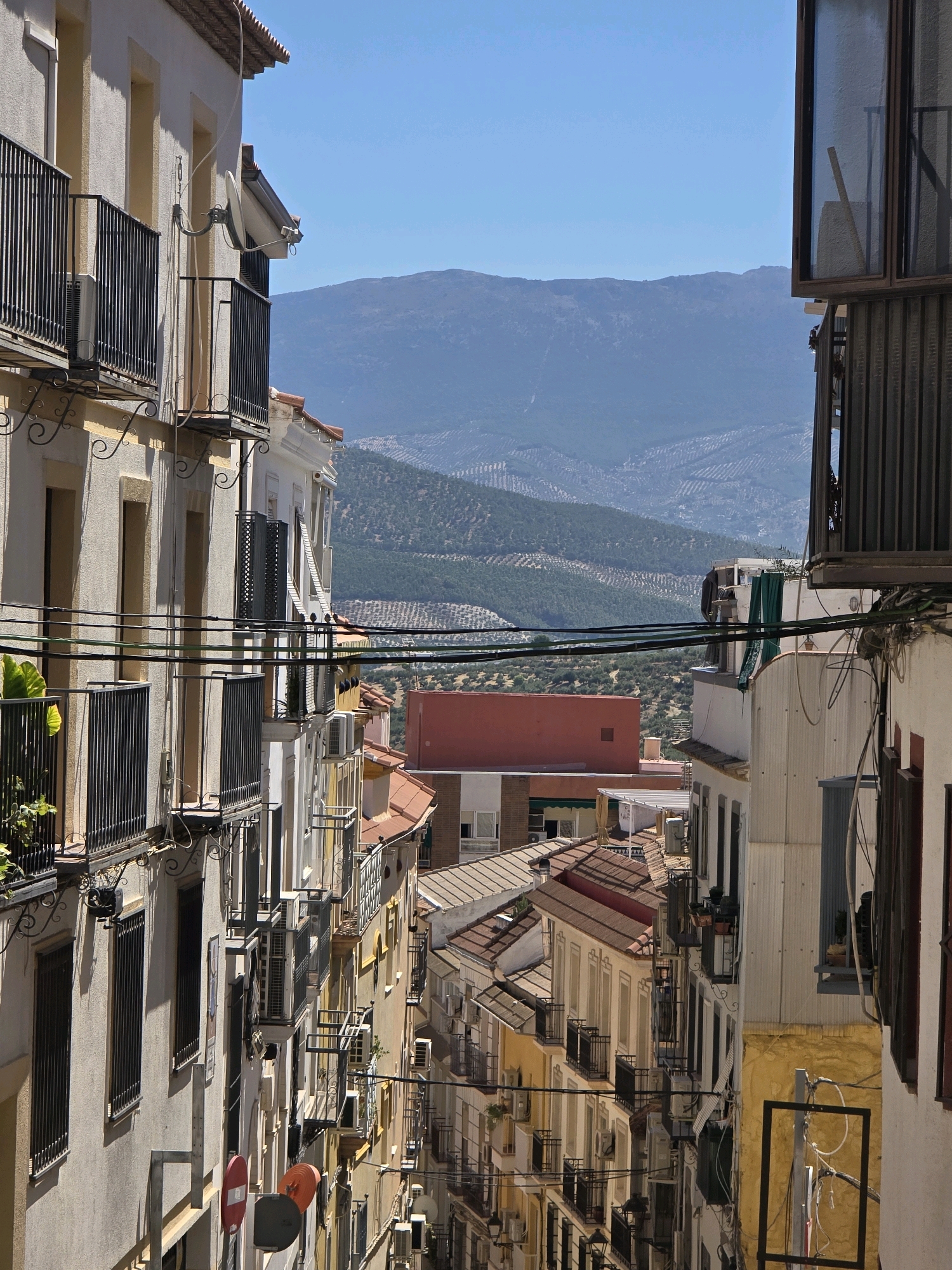 The image captures a narrow street in the old town of Jaén, Spain, looking down a steep incline towards distant mountains. The architecture is characterized by multi-story buildings with stucco facades, numerous balconies with wrought-iron railings, and terracotta-tiled roofs. Several air conditioning units are visible mounted on the walls and balconies. Electrical wires crisscross the scene overhead. In the foreground, two circular signs, one red and white and another gray, are mounted on a pole. The landscape beyond the urban area features rolling hills covered in what appear to be olive groves, leading up to a hazy, blue mountain range under a clear sky. The overall impression is of a dense, historic urban environment with a strong connection to the surrounding agricultural landscape.