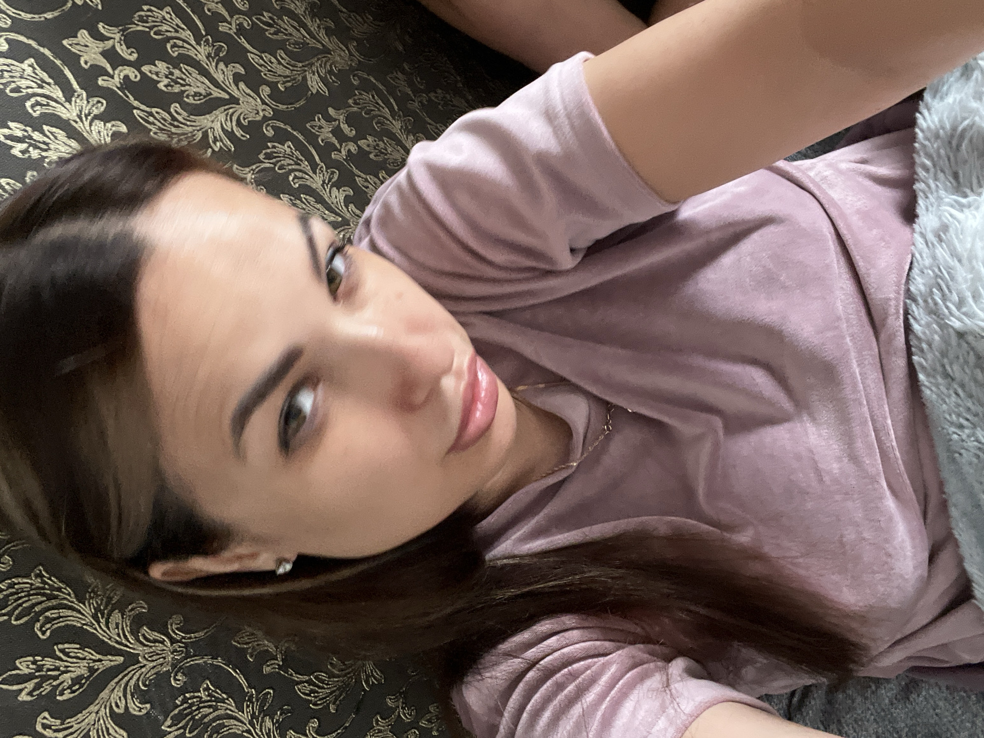 A selfie of a young woman with long, dark brown hair and brown eyes is shown. She is wearing a light pink, velvety t-shirt and a thin silver necklace. Her head is tilted to her left, and she is looking directly at the camera with a slight pout. The background consists of dark wallpaper with a gold floral pattern and what appears to be a textured sofa or blanket. The lighting suggests an indoor setting.