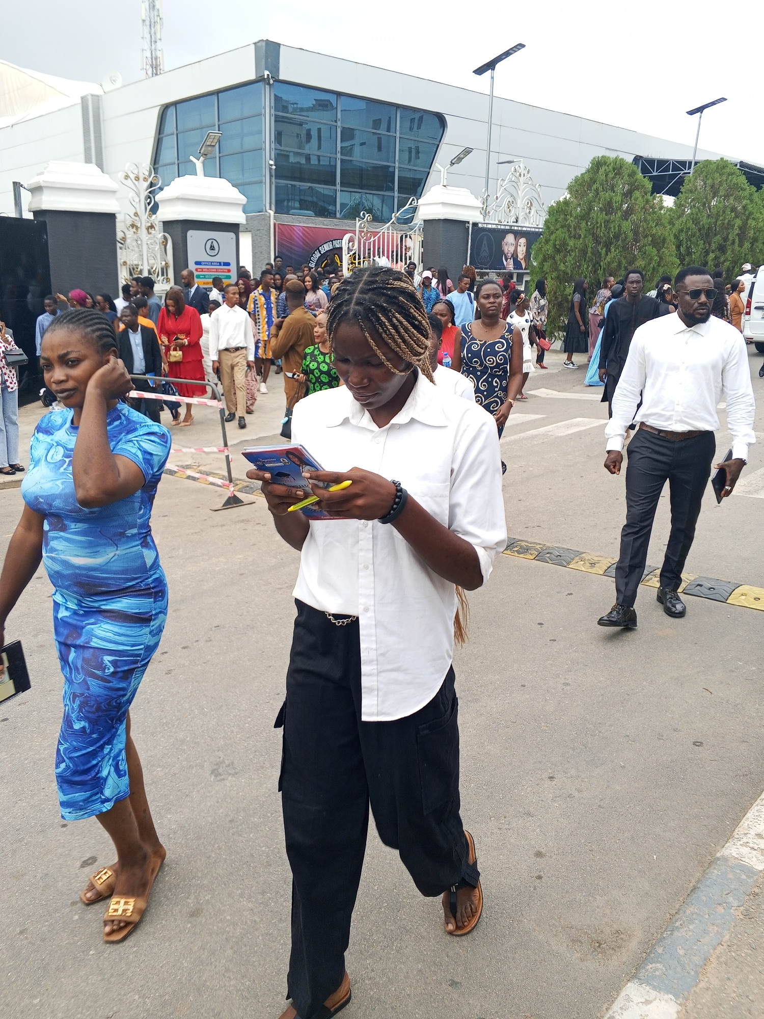 The image captures an outdoor scene in front of a large, modern building with a glass facade, likely in Abuja, Nigeria. A crowd of people, appearing to be attending an event or gathering, are walking on a paved road. In the foreground, a woman in a blue, patterned dress walks towards the left, while another person in a white collared shirt and black pants, holding a booklet and a pen, walks towards the right, seemingly engrossed in the booklet. Other individuals in the background are dressed in various casual and formal attire. The environment suggests a daytime event, with some overhead structures and trees visible in the distance.