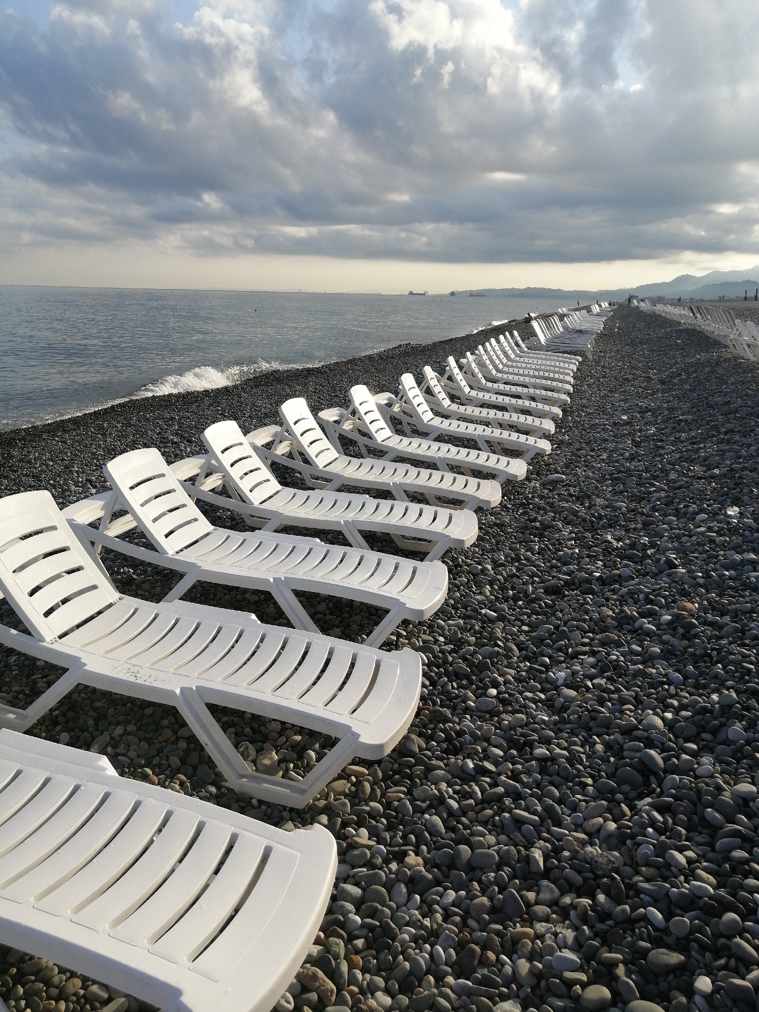 A long perspective of empty, white plastic lounge chairs on a pebble beach is shown. The chairs are neatly arranged in rows, facing the sea. The sea is calm and grey, with small waves lapping at the shore. The sky is overcast with dramatic clouds. In the distance, there are hills and mountains. The scene appears to be located on the Batumi embankment in Georgia, a popular tourist destination. The image evokes a sense of solitude and tranquility, with the abandoned chairs hinting at a recent or upcoming influx of visitors.