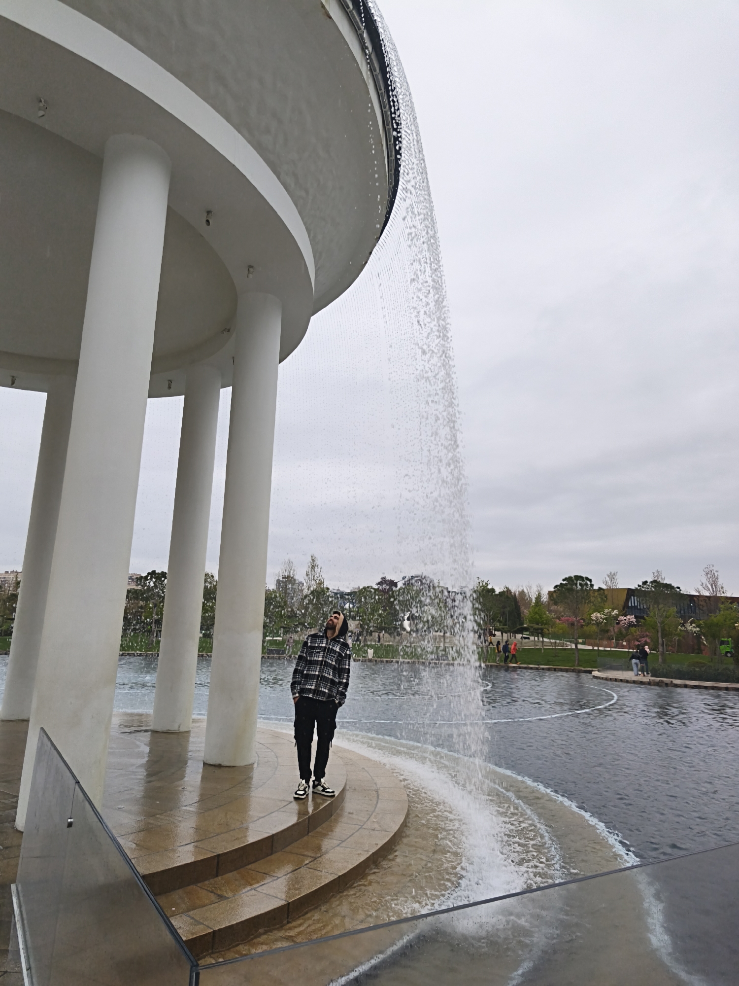 A man stands within the "Infinity" water fountain in Krasnodar, Russia. The fountain features a circular, white, dome-like structure supported by multiple white columns. Water cascades down the exterior of the dome, creating a curtain. The man is standing on the tiled platform of the fountain. He is wearing a plaid jacket, dark pants, and shoes. He is looking upwards towards the fountain's structure. The background includes a body of water surrounding the fountain, trees, other landscaping, and people walking in the distance. The sky is overcast. A glass barrier is visible in the foreground.