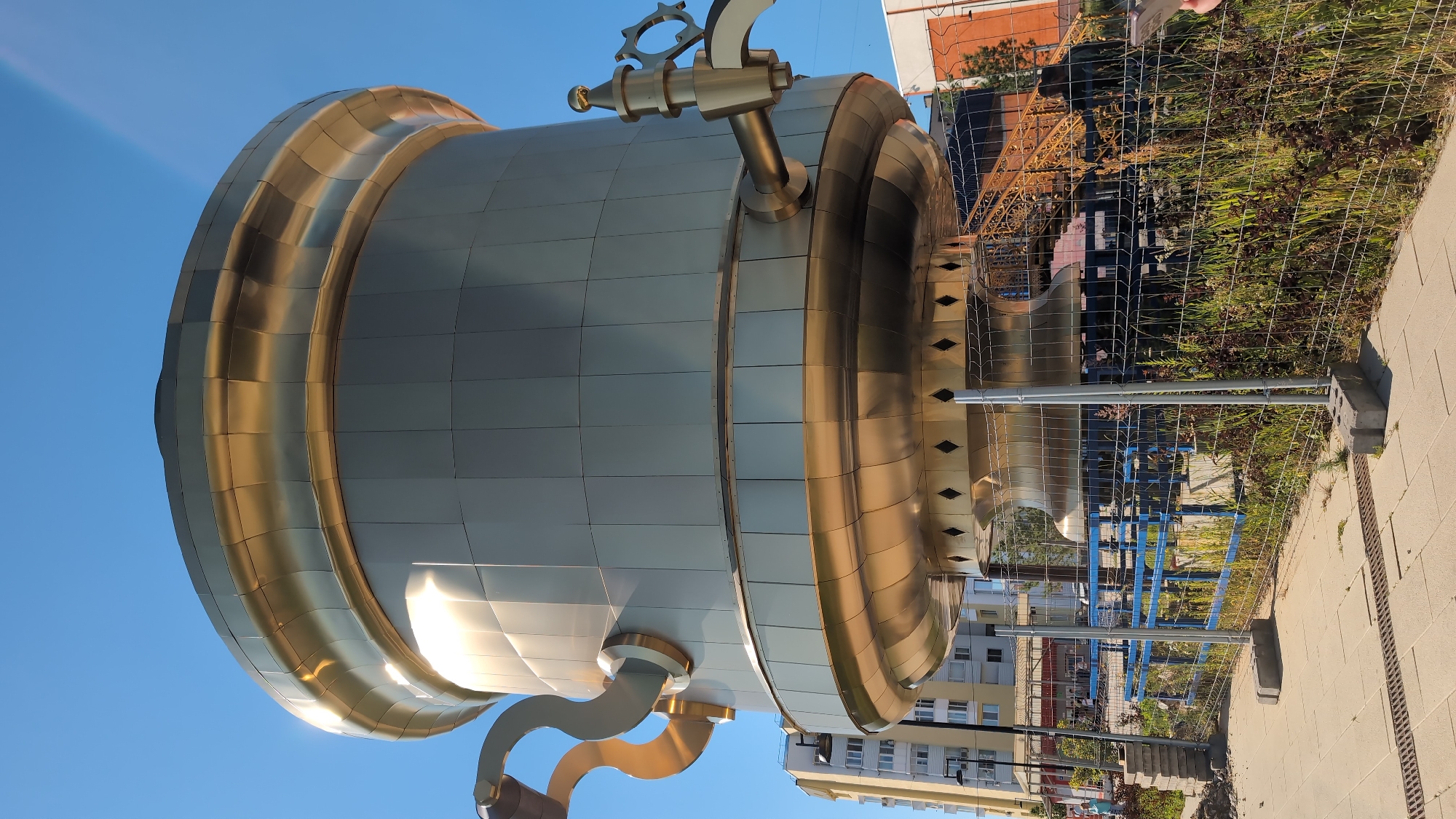 This media file features a large, metallic sculpture resembling a kettle, predominantly in shades of silver and gold. The sculpture is oriented vertically, with its base visible at the bottom of the frame. It has a rounded, bulbous body with distinct horizontal bands of gold. A golden spout-like appendage extends from the lower left side of the sculpture. On the upper portion of the sculpture, a golden mechanical element, possibly a valve or a decorative gear, is attached. The sculpture is situated outdoors, with a clear blue sky dominating the background.
In the background, beyond a wire mesh fence, there are buildings, trees with autumn foliage, and some construction materials or equipment, including blue scaffolding or supports and yellow girders, suggesting ongoing construction or maintenance in the area. A paved walkway is visible to the right of the fence, with concrete blocks serving as anchors for the fencing.
The lighting suggests it is daytime, with bright sunlight casting reflections on the metallic surfaces of the sculpture, indicating clear weather. The scene appears to be in a public outdoor space, possibly a park or a plaza undergoing development or renovation. No people are clearly discernible in the frame, but the presence of construction elements implies human activity in the vicinity. The location context provided is Bor, Russia. There is no visible text in the image.