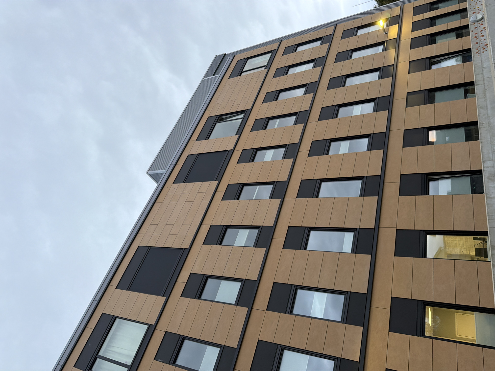The image shows the exterior of a modern building in the city of Harstad, Norway. The building features a facade of light brown, wood-like panels interspersed with dark vertical and horizontal elements. Numerous windows are visible, many with dark frames. The building is angled from a low perspective, with the sky taking up a significant portion of the upper frame. The sky is overcast with a pale, cloudy appearance. A street lamp is visible on the right side of the building, illuminated with a yellow light. The lower part of the image shows a concrete barrier or wall, partially obscuring the base of the building. The overall impression is one of contemporary architecture under a typically Nordic cloudy sky.