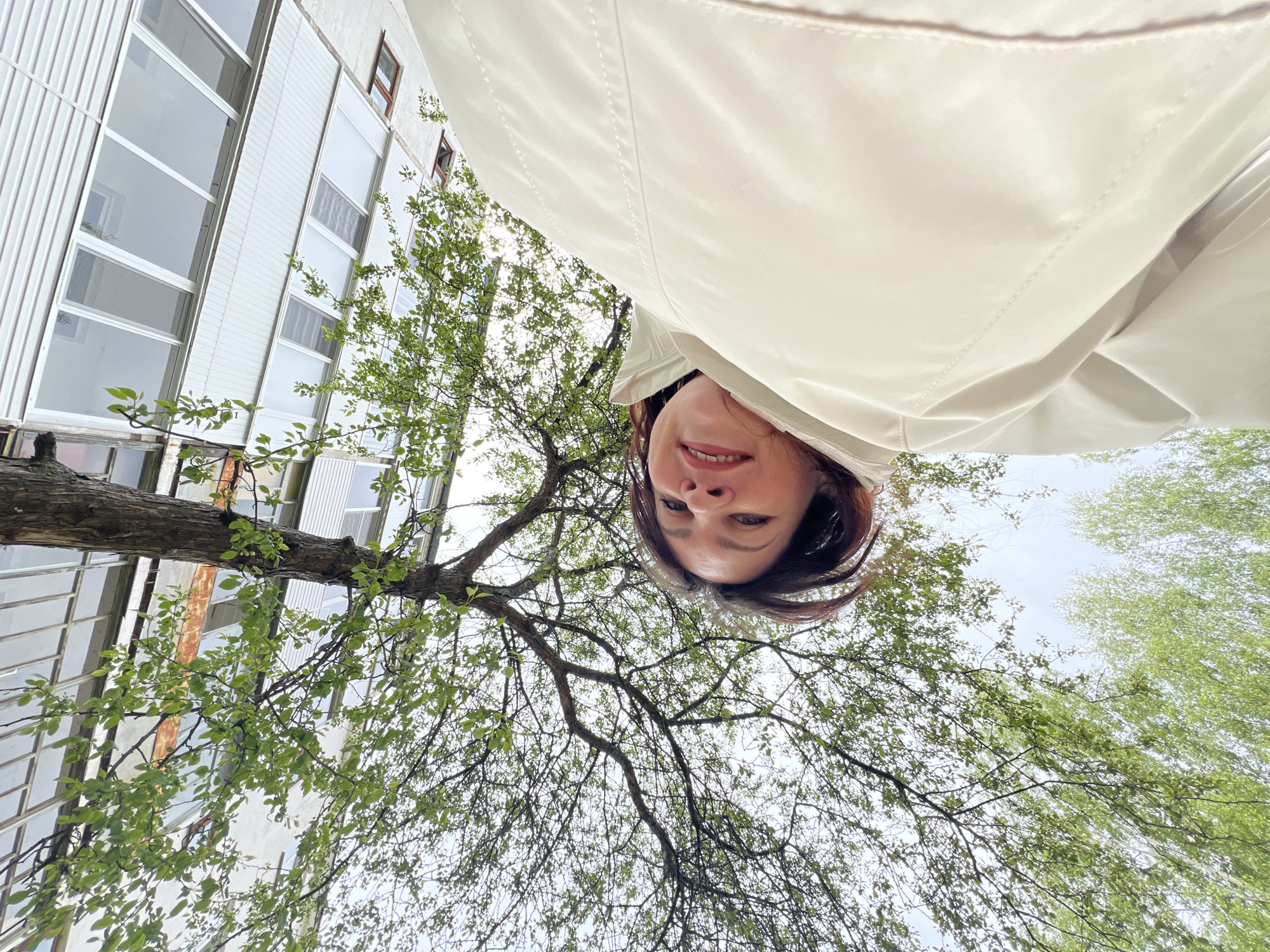 A low-angle shot looking upward captures a woman's face, a tree with green leaves, and a building. The woman, likely holding the camera, has dark hair and is partially obscured by what appears to be a light-colored jacket. Above her and the tree, the sky is visible. To the left, a building with multiple windows can be seen. The location is possibly in the city of Noviy, Russia.
