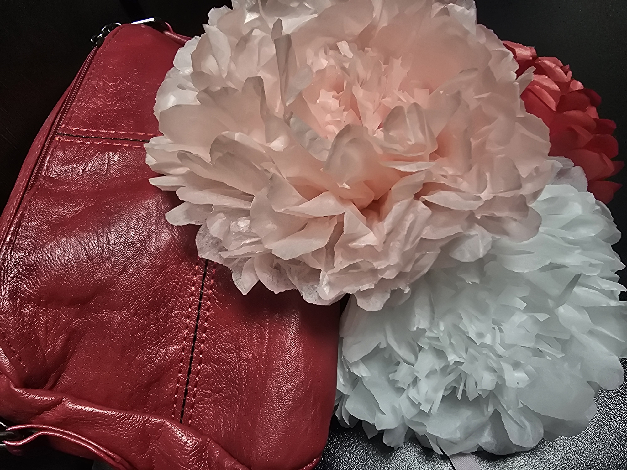 A red leather handbag is partially visible at the top of the frame, with its zippered closure and stitching clearly depicted. In front of the handbag, three large, fluffy paper pom-poms are arranged. The pom-poms are in shades of white, pale pink, and a brighter reddish-pink. The background is dark and out of focus. The scene appears to be a still life composition, possibly set up for a product photograph or a decorative arrangement. No specific location in the city of Unknown, Unknown is identifiable from the image.