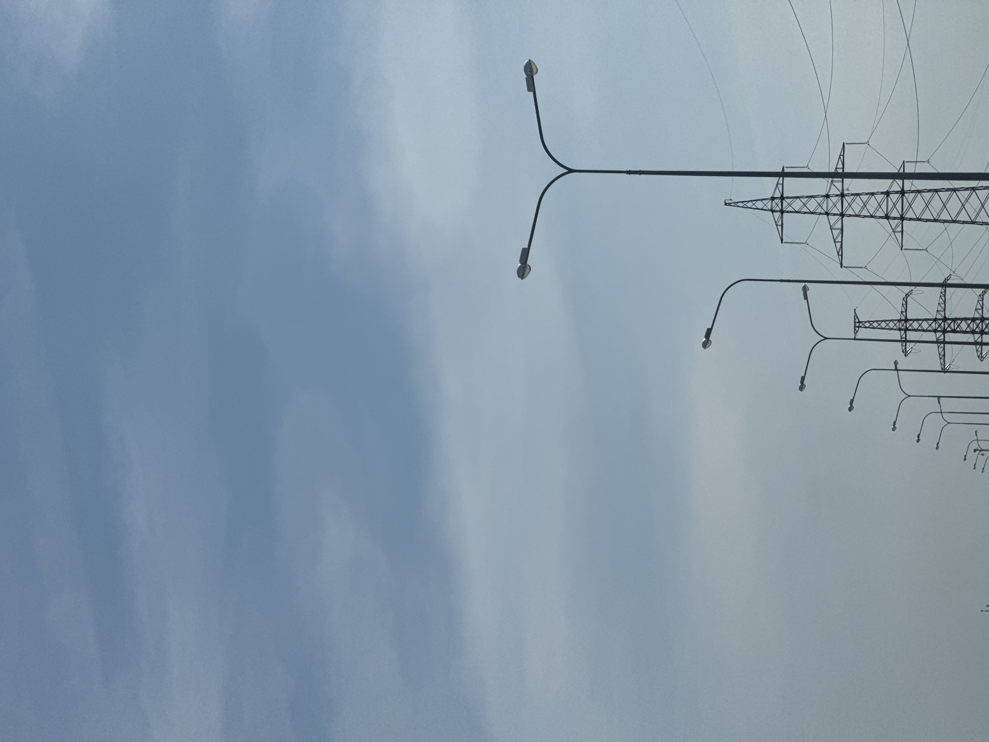 The image depicts a row of streetlights and power lines against a light blue sky. The lights are situated along a road or highway, receding into the distance. The power lines, supported by metal towers, run parallel to the streetlights. The image is rotated 90 degrees counter-clockwise. The city of Noviy, Russia is not verifiable from this image.
