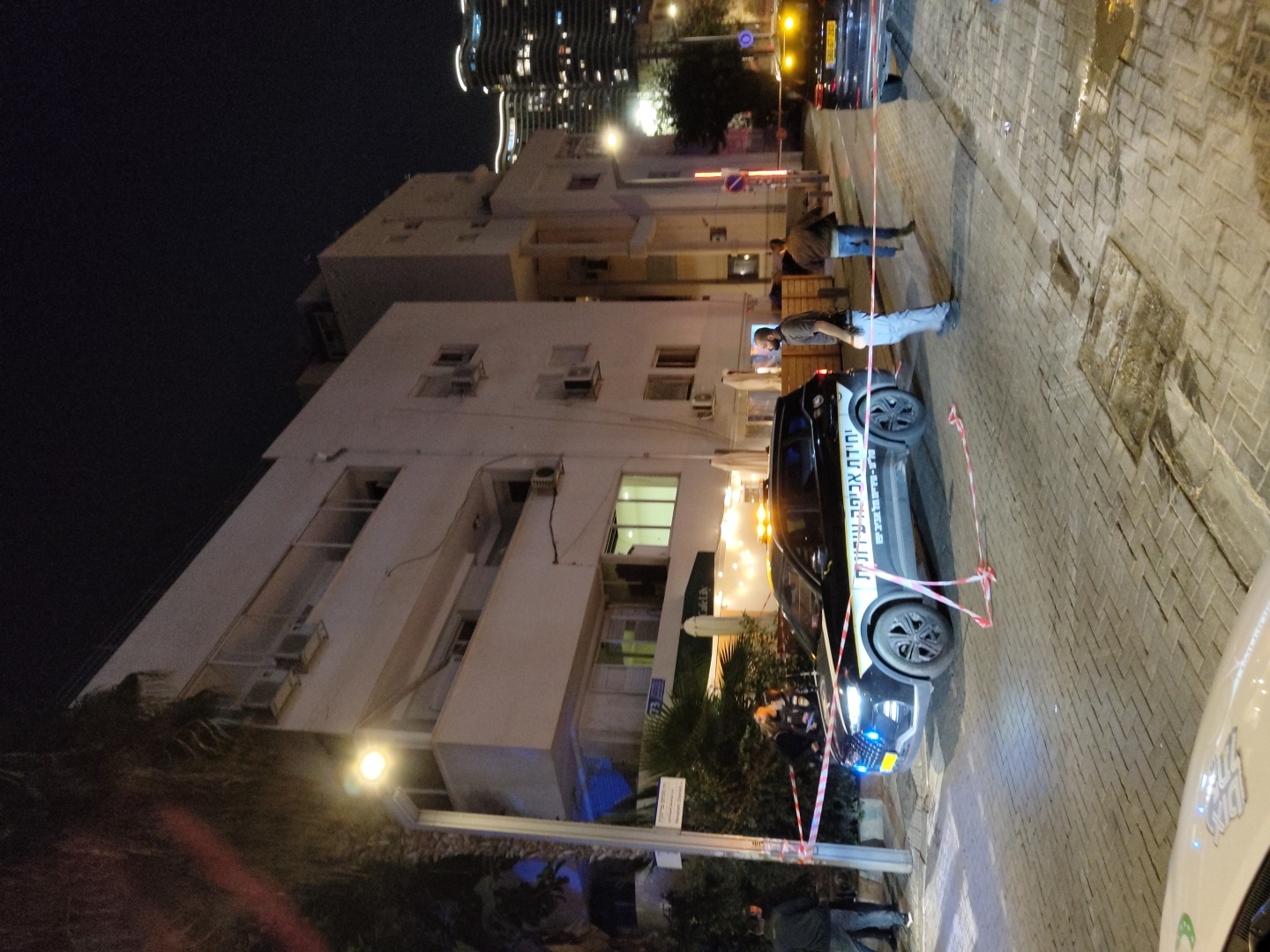 The image depicts a nighttime urban scene on a paved street in Tel-Aviv, Israel, indicating a cordoned-off area. A black SUV, identified as a "Municipal Enforcement Unit" ("יחידת אכיפה עירונית") vehicle with white and black markings and amber flashing lights, is centrally positioned. Red and white striped barrier tape is strung around the SUV and extends across the cobblestone street, delineating a restricted zone.
Multiple individuals are present: a male in a dark shirt and light blue jeans stands near the left side of the SUV, facing right. Another male in a brown jacket and blue jeans walks away from the scene on the right side of the street, heading towards a second dark SUV partially visible in the background. Additional figures are dimly visible near the front of the enforcement vehicle.
The background features a white, multi-story building, identified by a sign as "Cafe Lily" and bearing the street number "73." The building exhibits balconies, windows, and exterior air conditioning units. Streetlights illuminate the scene, casting shadows. Palm trees are visible on the left. In the distance, illuminated high-rise buildings of the city skyline are visible against the dark night sky. The foreground shows part of a white vehicle's hood.