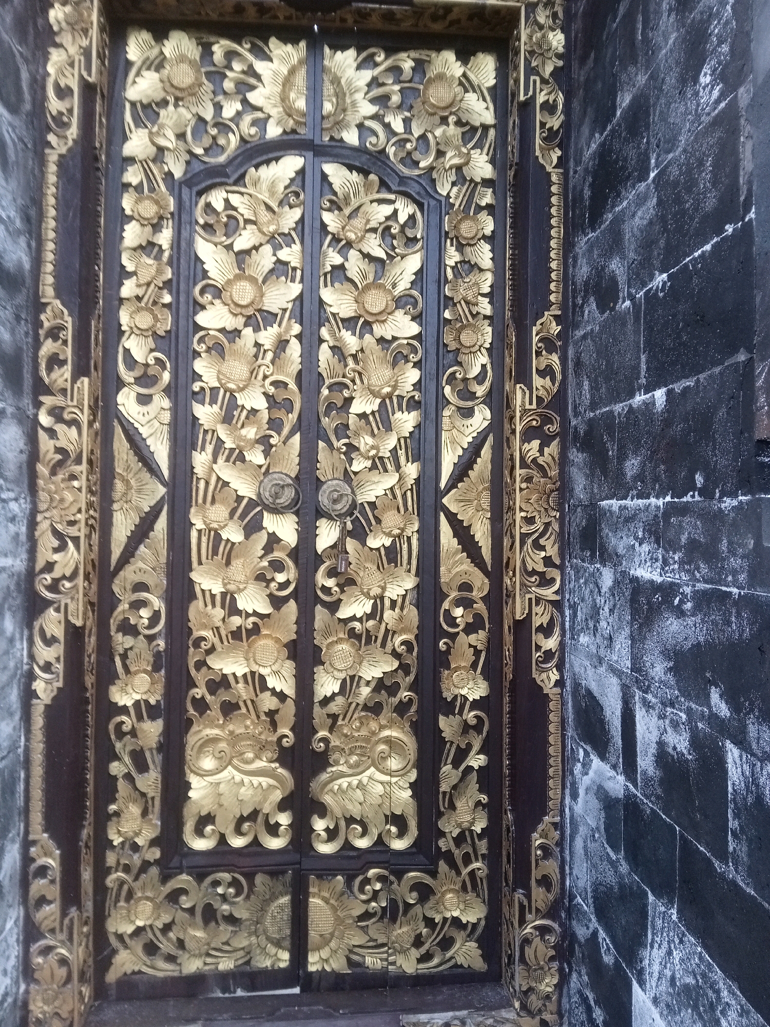 A detailed, ornate, golden carved wooden door with intricate floral patterns stands against a dark, weathered brick wall. The door is divided into two panels, each adorned with a symmetrical arrangement of large flowers with multiple petals and smaller blossoms interspersed with leaves and swirling tendrils. The gold carving contrasts sharply with the dark wood of the door and the black of the brick wall. The brick wall appears rough and aged, with streaks of white or light gray weathering. The scene is lit from the front, casting subtle shadows that highlight the dimensionality of the carvings. There are two circular door handles, one on each panel, made of a dark metal, and a key is visible in the lock of the right-hand door handle. The perspective is a slightly angled, medium shot, focusing on the texture and detail of the door and its immediate surroundings. The background beyond the brick wall is not visible.