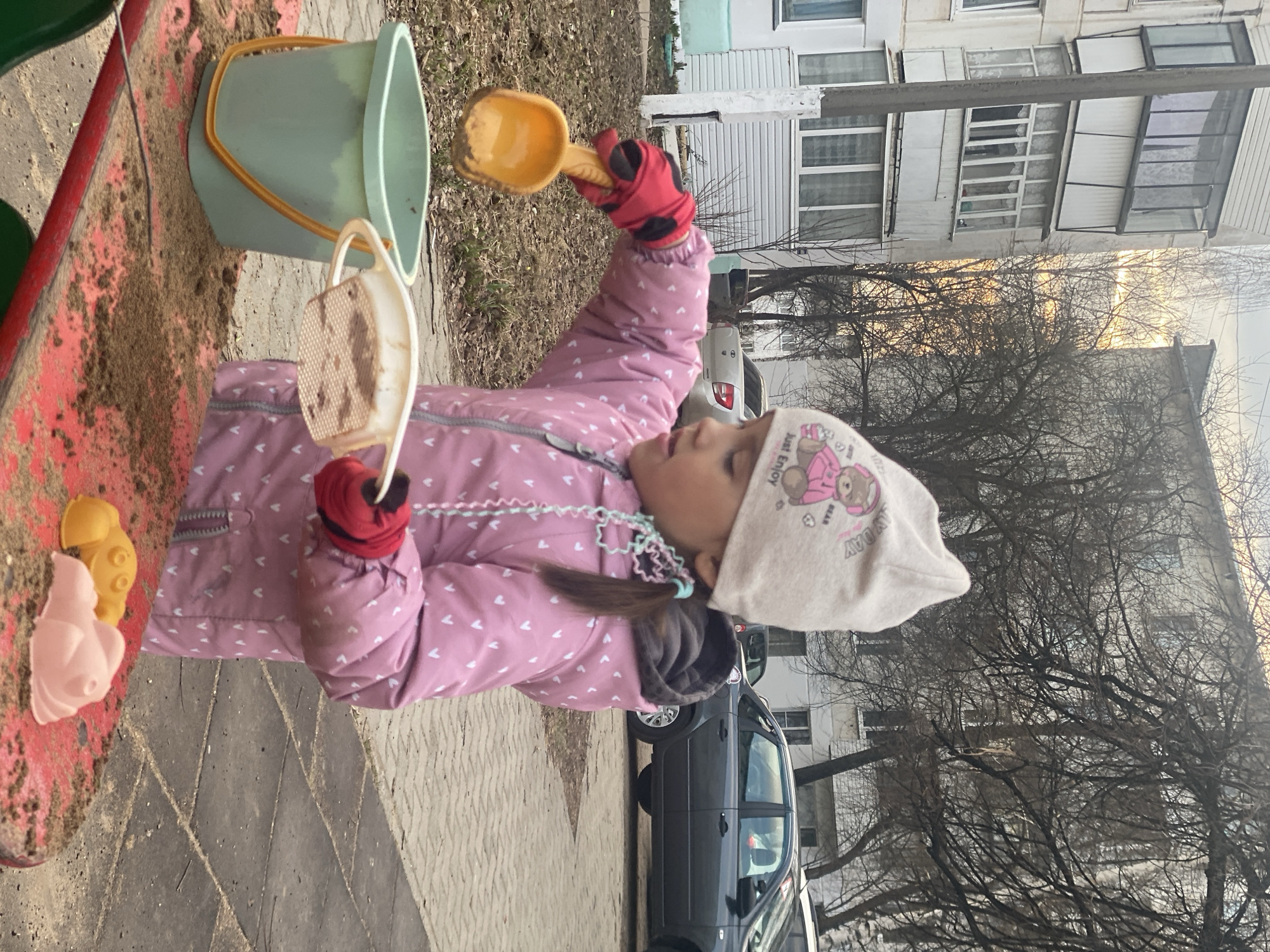 A young child is captured playing in a sandbox outdoors. She is dressed in a pink puffer jacket patterned with white hearts, red gloves with black polka dots, and a light-colored beanie. The beanie displays the text "JUST ENJOY" and "BEAR" next to a cartoon bear illustration. She holds a yellow sand shovel aloft in her right hand and a white sand sifter in her left, actively engaged with the sand. A light green bucket with a yellow handle sits near the red edge of the sandbox, containing some sand. Nearby, pink and yellow sand molds, resembling a seashell and a star or flower, rest on the sandbox edge.
The child is standing on a paved area, which transitions into a street where a dark grey car is parked. In the background, a multi-story residential building with numerous windows is visible. The scene is framed by several bare trees, suggesting late autumn, winter, or early spring weather. The sky is bright but appears overcast, indicating it is daytime, possibly late afternoon due to a subtle golden light filtering through the trees. The child appears focused on her play. This scene takes place in an urban or suburban setting, specifically in Chaikovsky, Russia.