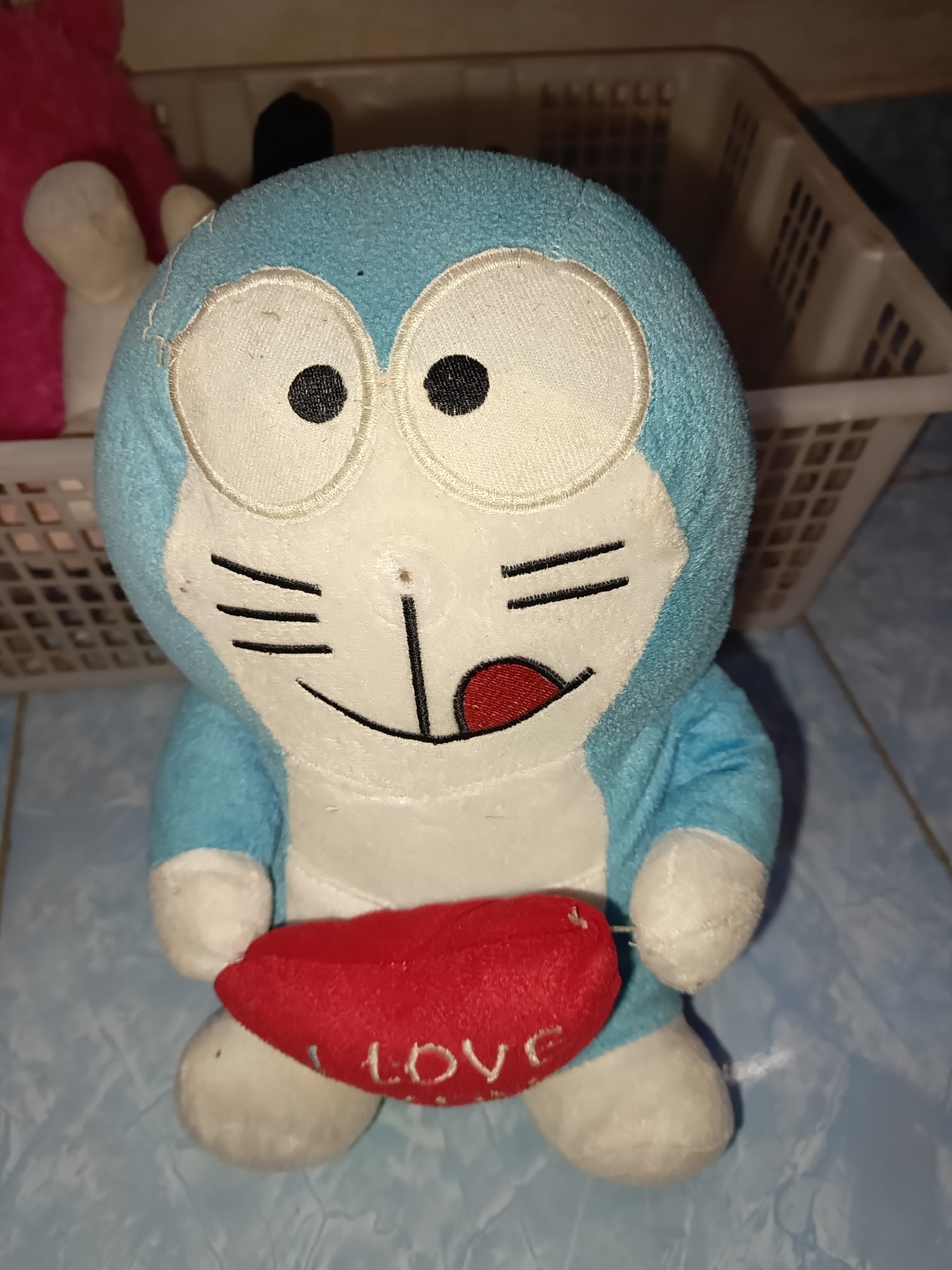 A stuffed toy resembling the character Doraemon is positioned centrally in the frame. The toy is blue and white, with large black and white eyes, a black nose, and a red tongue sticking out. It is holding a red, heart-shaped object with the word "LOVE" embroidered on it in white. The toy is sitting on a light blue, tiled surface. In the background, a white plastic basket is visible, containing other items, one of which appears to be pink. The overall setting suggests an indoor environment, likely a room in a home in Terbanggi Marga, Indonesia.