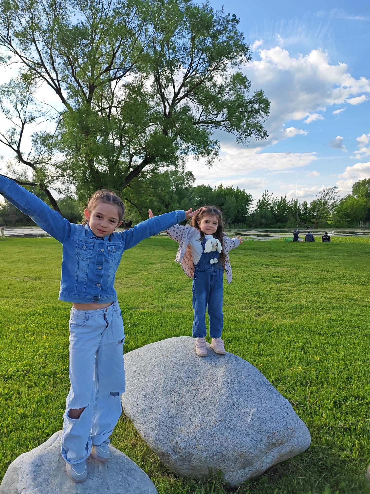 Two young girls stand on large rocks in a grassy field. The girl on the left wears a denim jacket and light blue ripped jeans, with her arms outstretched. The girl on the right is in denim overalls with a patterned sweater, also with outstretched arms. In the background, there's a large tree and several people sitting near a body of water. The sky is blue with scattered clouds. The scene is in Bronnitsy, Russia.