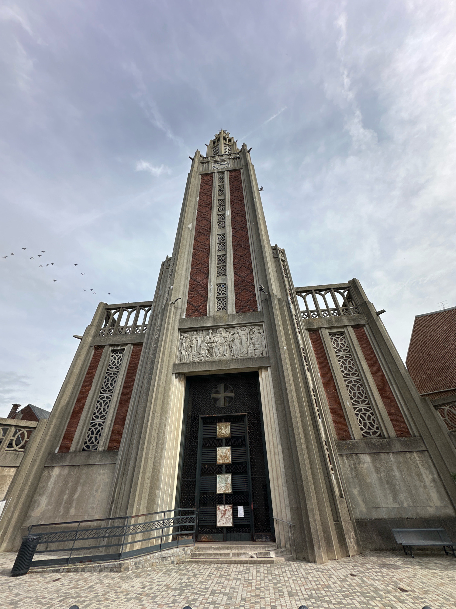 The photograph captures the imposing facade of the Notre-Dame church in Roye, France. The shot is taken from a low angle, looking upwards towards the tall, art deco-style tower which dominates the center of the image. The tower features a blend of concrete and red brick, with intricate patterned windows and decorative relief sculptures above the entrance. A flock of birds is visible in the sky to the left of the tower. The ground in front of the church is paved with cobblestones, and a dark metal railing and a bench are present on the right side of the frame. The sky is overcast with scattered clouds.