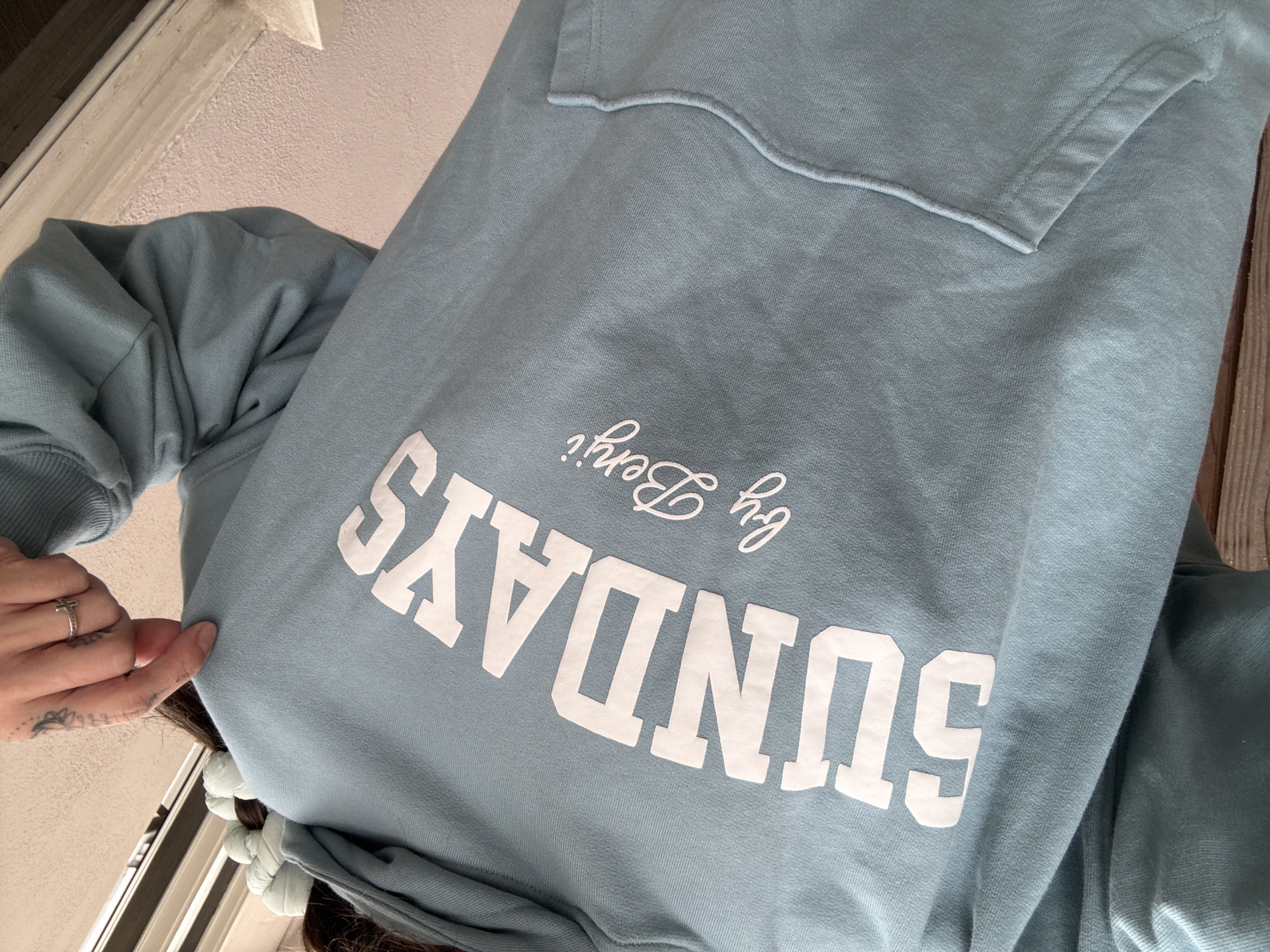 A close-up, slightly angled shot focuses on a light blue sweatshirt. The word "SUNDAYS" is prominently printed in white, block letters on the garment. Above "SUNDAYS," the text "by Berri" is written in a cursive font, also in white. The sweatshirt appears to have a pocket, visible at the top of the image. A hand with a silver ring and some tattoo ink on the wrist is visible on the left side of the image, holding onto the sweatshirt. The background is out of focus, but shows a white wall and what seems to be part of a wooden structure. It is not possible to determine the location in Seal Beach, United States of America from this image.