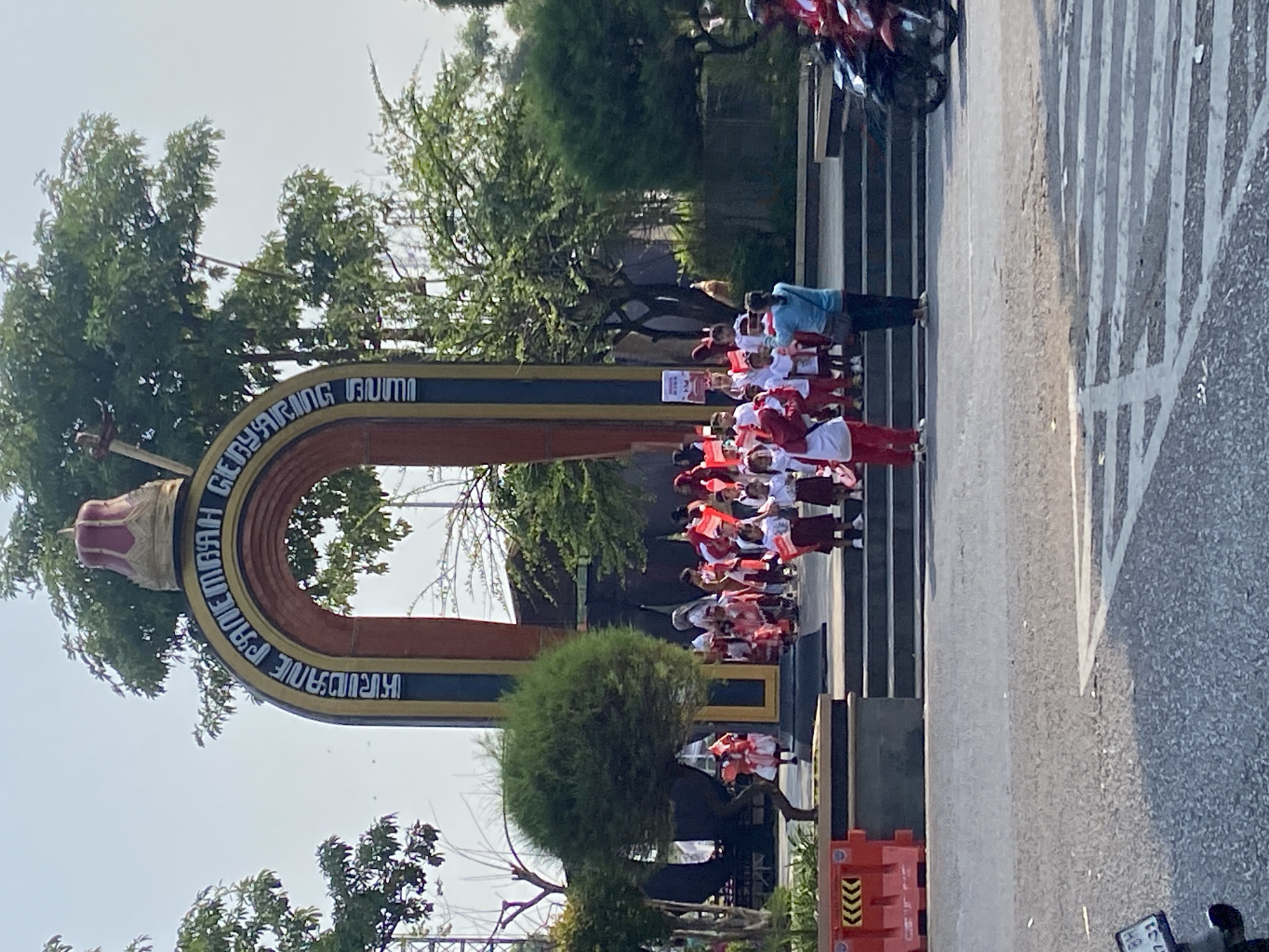 In front of a large arched monument with text in Indonesian, a group of children in school uniforms and carrying small Indonesian flags are gathered on a set of stairs. The monument is decorated with red and gold colors and has a crown-like structure at its apex. The children appear to be participating in a parade or ceremony, with some adults present to supervise. The scene is set outdoors, with trees and buildings visible in the background. The photograph appears to have been taken on a sunny day. The monument is located in the city of Kecamatan Pati, Indonesia.