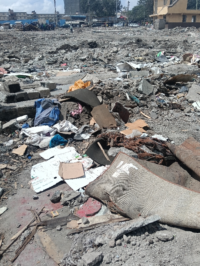 The image depicts an expansive urban demolition site in Nairobi, Kenya, under bright daylight with a hazy sky. The ground, from foreground to midground, is uniformly covered with grey rubble, broken concrete, rebar, shattered construction materials, and loose dirt, indicating recent structural clearance.
In the foreground, specific debris elements include a folded, soiled, light-colored woven mat with a faded pattern, fragmented cardboard pieces, scattered white paper, various discarded textiles (notably blue, white, and pink fabrics), irregular dark synthetic material fragments, and corroded metal. A blue backpack is situated amidst concrete blocks in the lower left. Patches of reddish discoloration stain the ground surface.
The midground continues the rubble field, forming uneven mounds of concrete and earth.
The background presents an urban periphery. To the far left, a group estimated at 15-20 individuals is clustered near a large mound of construction debris. A solitary individual walks across the distant cleared area. Further left, a low-rise building with a blue roof is discernible, alongside multiple white vehicles. A dirt road or cleared pathway runs horizontally across the mid-distance, populated by vehicles. Beyond this, taller multi-story buildings are visible. To the right, a multi-story, yellow-painted building, partially demolished or structurally compromised, stands. Trees are visible in the far background. No active operations are in progress; the scene depicts a static, post-demolition landscape.