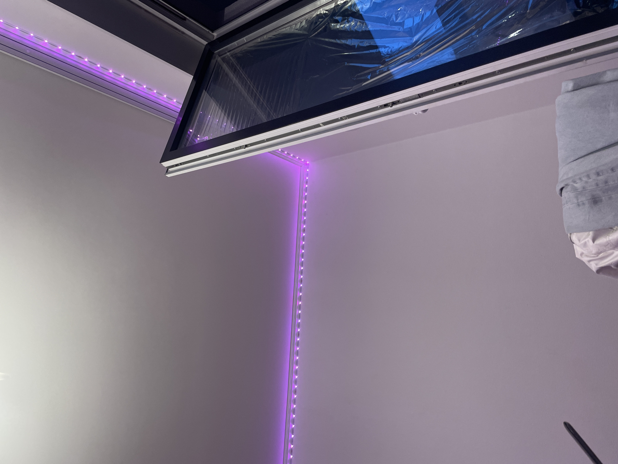 The image shows an interior scene with purple LED strip lights running along the top of the walls and ceiling. A window is partially open, revealing a dark interior with blue-tinted plastic sheeting. The lower portion of the image shows a stack of clothing, including a pair of light blue jeans and a white garment. There is a dark, thin object on the left side of the frame, possibly a stand or a pole. The scene is likely indoors, and there is no indication of its specific location within Irinovka, Russia, or any interaction between individuals.