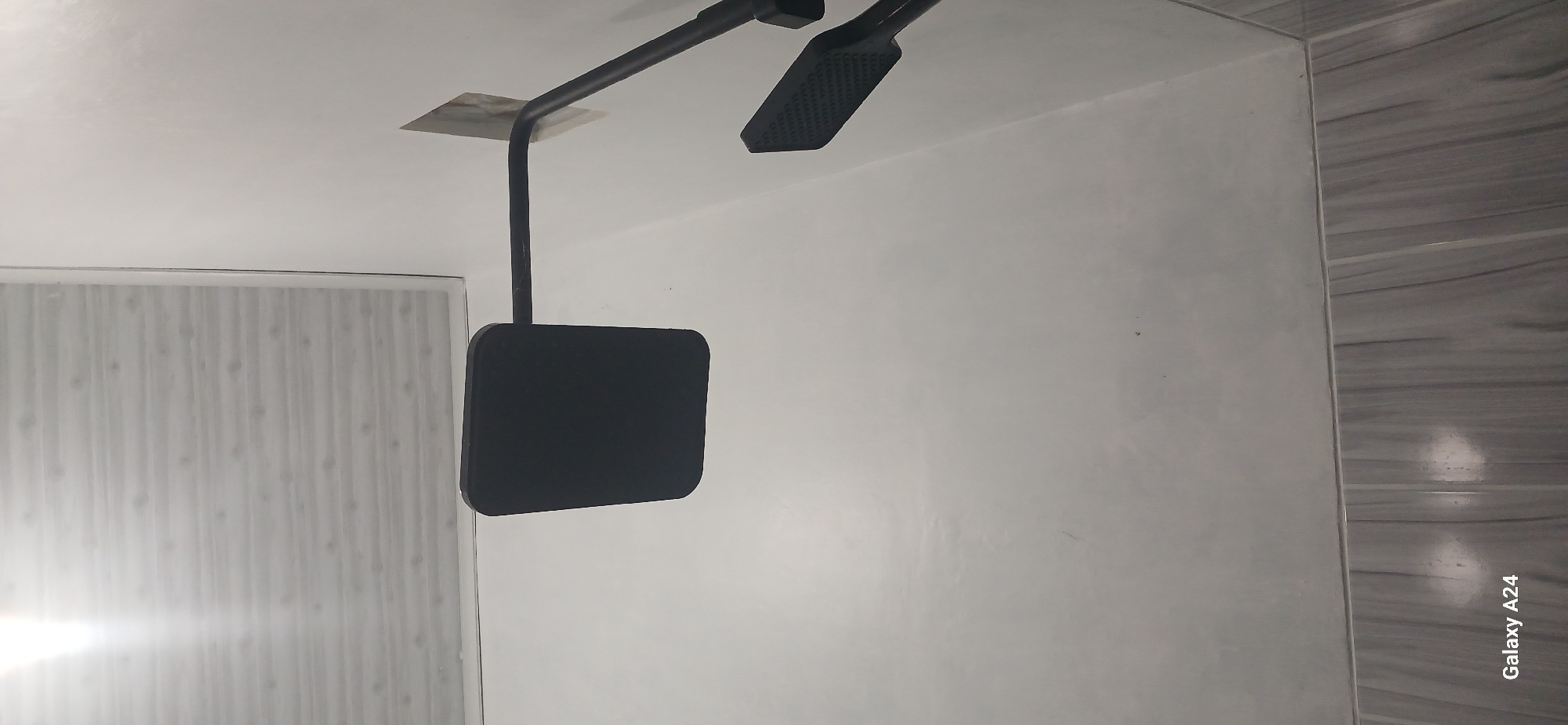 The image shows a modern shower head attached to a dark-colored pipe, mounted on a white wall. The shower head itself is rectangular with rounded corners and appears to be made of a dark material, possibly black. The pipe extends from the wall horizontally and then curves downwards. Below and to the right of the main shower head, another, smaller shower head is visible, also in a dark color and with a textured surface. The wall is painted white and has a slightly textured finish. The ceiling above is paneled with a wood-grain pattern, possibly white wood-effect panels. The floor, visible at the bottom of the image, is tiled with grey and white striped tiles, laid horizontally. The overall impression is of a contemporary bathroom. The image is a photograph taken from a slightly low angle, looking up at the shower fixture. The metadata "Galaxy A24" indicates the image was taken with a Samsung Galaxy A24 smartphone. There is no identifiable information within the image that suggests a specific location in Kuwarisan, Indonesia.
