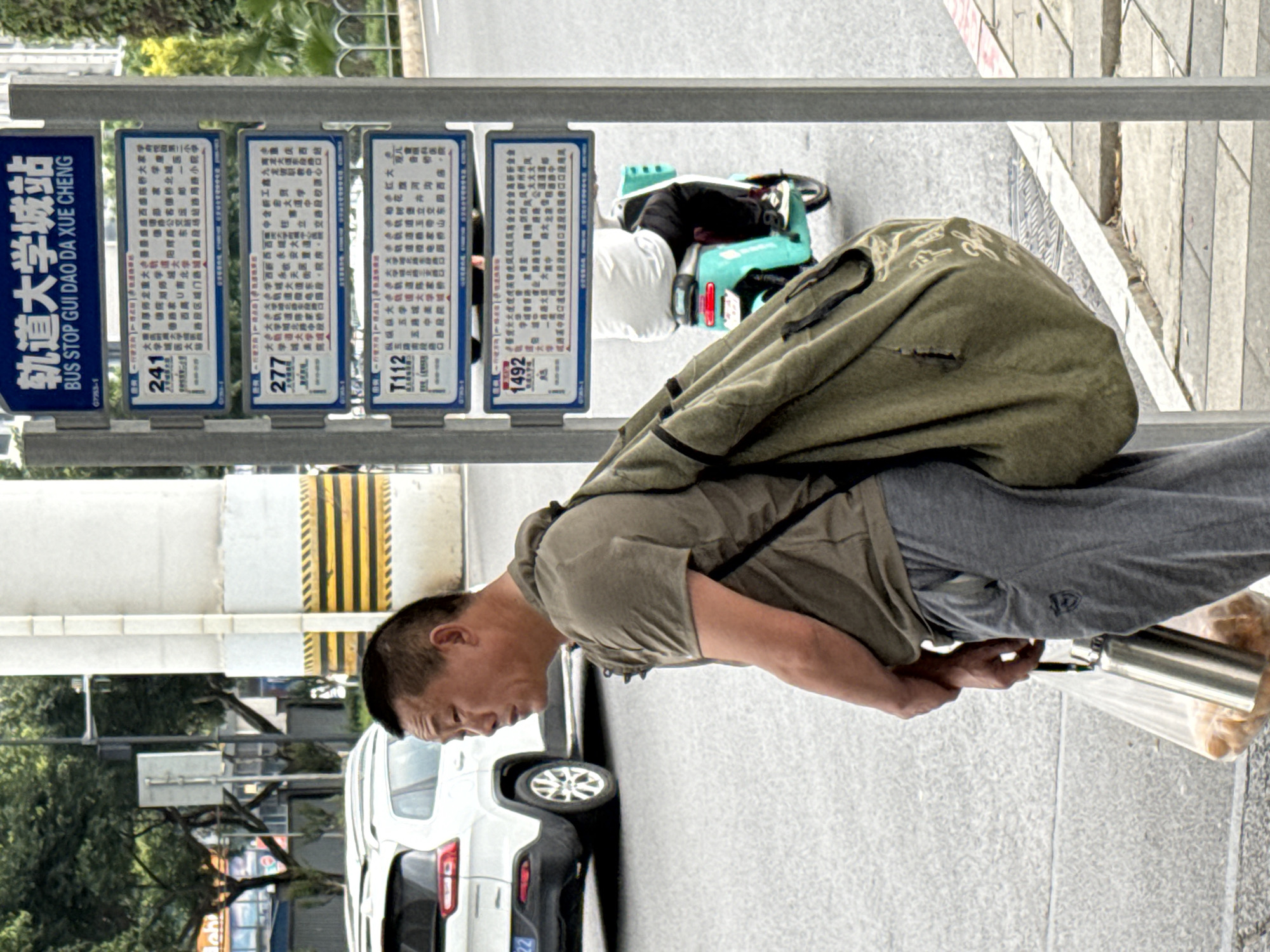 The image shows a man in Chongqing High-tech Zone, China, seemingly fixing something on the ground with a metal container and plastic bag nearby. He is wearing a green shirt, gray pants, and a large bag over his shoulder. The scene is set at a bus stop, with signs for various bus routes (241, 277, T112, and 1492) displayed in Chinese, along with the station name "轨道大学城站" (Gui Dao Da Xue Cheng - University City Railway Station). A white car is partially visible behind the man. The environment includes a sidewalk and street with some greenery visible in the background.