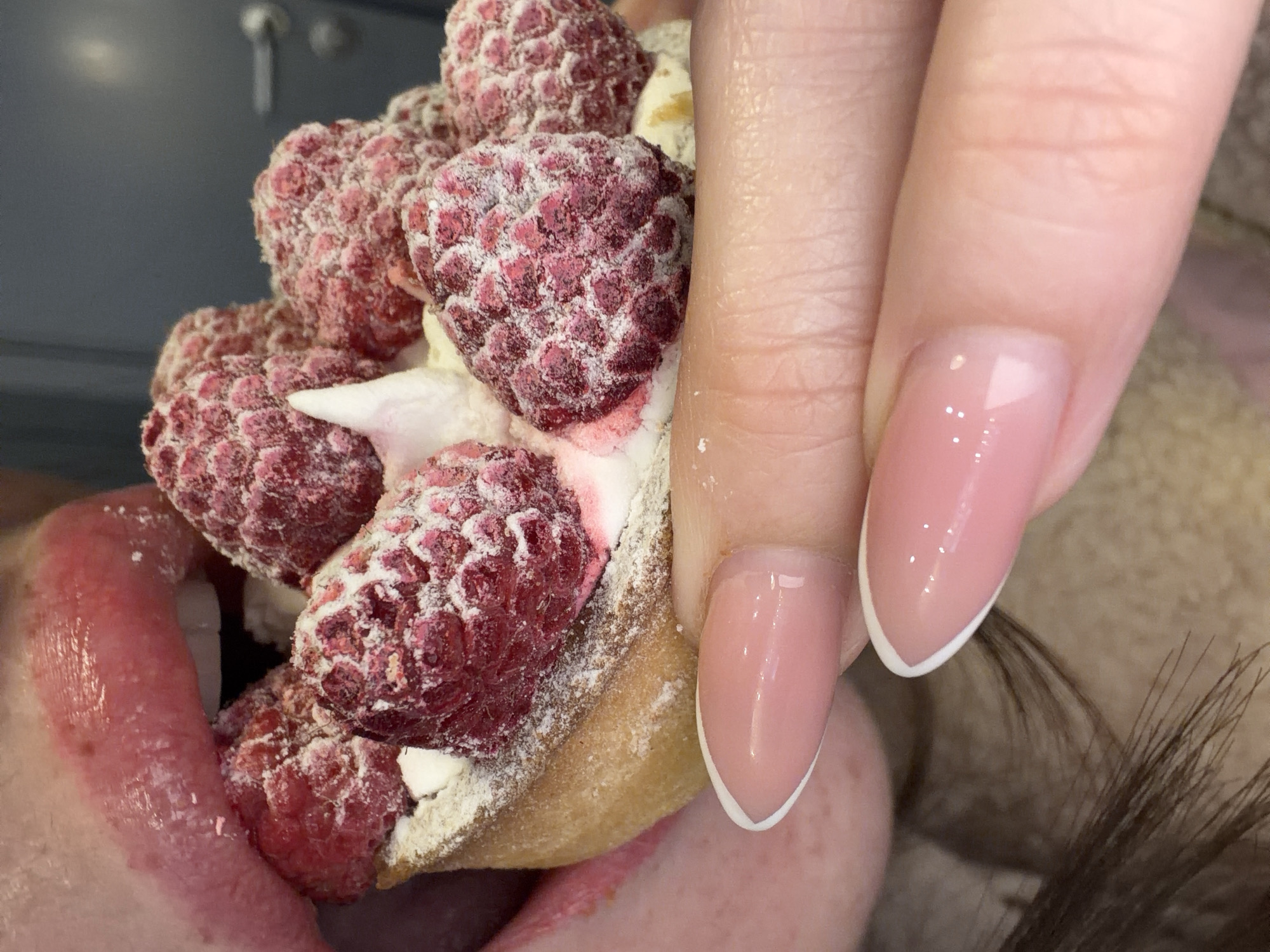 The image presents a close-up, high-angle view of an individual in the act of consuming a raspberry tart.
**People:** The lower portion of a person's face is visible on the left, featuring full, glossy, reddish-pink lips, slightly parted to expose white teeth and the tip of a tongue. The tongue is extended towards the dessert. A right hand, with fingers holding the tart, occupies the lower central and right areas. The fingernails are manicured in a French tip style, displaying a light pink base with distinct white tips. Minimal dark hair or fabric is visible on the far lower-left side.
**Subjects/Objects:** The central subject is a round raspberry tart. It features a golden-brown pastry shell filled with a white, creamy substance, abundantly topped with multiple red raspberries. The raspberries are coated with a fine white powder, consistent with powdered sugar, giving them a frosted appearance. A prominent peak of white cream is visible amongst the berries. The tart is held firmly between the fingers of the right hand. The background consists of an out-of-focus, solid dark grey surface, possibly a wall or door, with a metallic silver door handle visible on the right side.
**Actions/Interactions:** The person is actively engaging with the dessert. The tongue is positioned as if licking or tasting the raspberries on the tart, which is held stable by the hand.
**Environment:** The background is generic and out of focus, indicating an indoor setting. No specific architectural features, landmarks, or identifying signs are present within the image to determine a precise location within the city of Saint Petersburg, Russia.