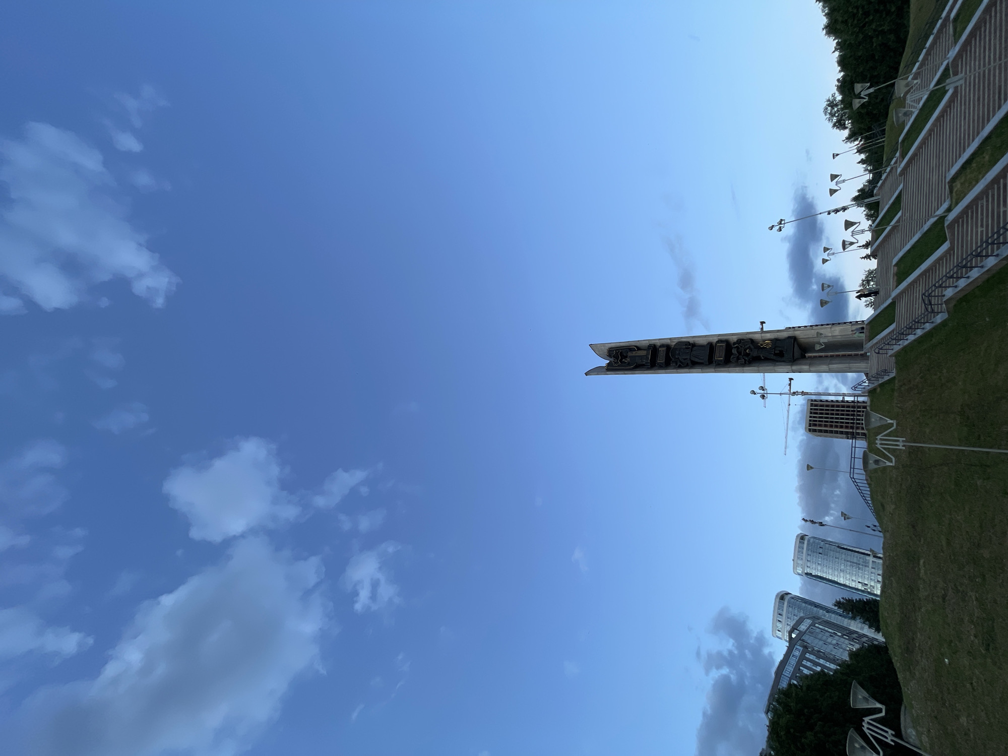 The image, oriented correctly, captures a contemporary urban landscape under a vast, clear blue sky dotted with scattered white and grey clouds. The sky fills approximately two-thirds of the frame. On the right side, a tall, narrow monument or sign stands prominently, featuring dark, stylized lettering that appears to spell "SCULPTURE" or a similar word. Behind and to the right of this monument, several modern high-rise buildings with reflective glass facades are visible, with at least one building under construction, indicated by a visible crane.
In the foreground and extending upwards along the right, there is a neatly maintained grassy slope traversed by concrete steps or terraces. Multiple path lighting fixtures, varying in design from single-lamp poles to multi-directional fixtures, are placed along the slope and adjacent areas. Clusters of green trees are visible at the upper right edge of the image and interspersed among the buildings. The lighting suggests a bright day, possibly late afternoon or early evening, with soft shadows. The overall scene combines urban development with natural elements and appears to be a public park or plaza area.
