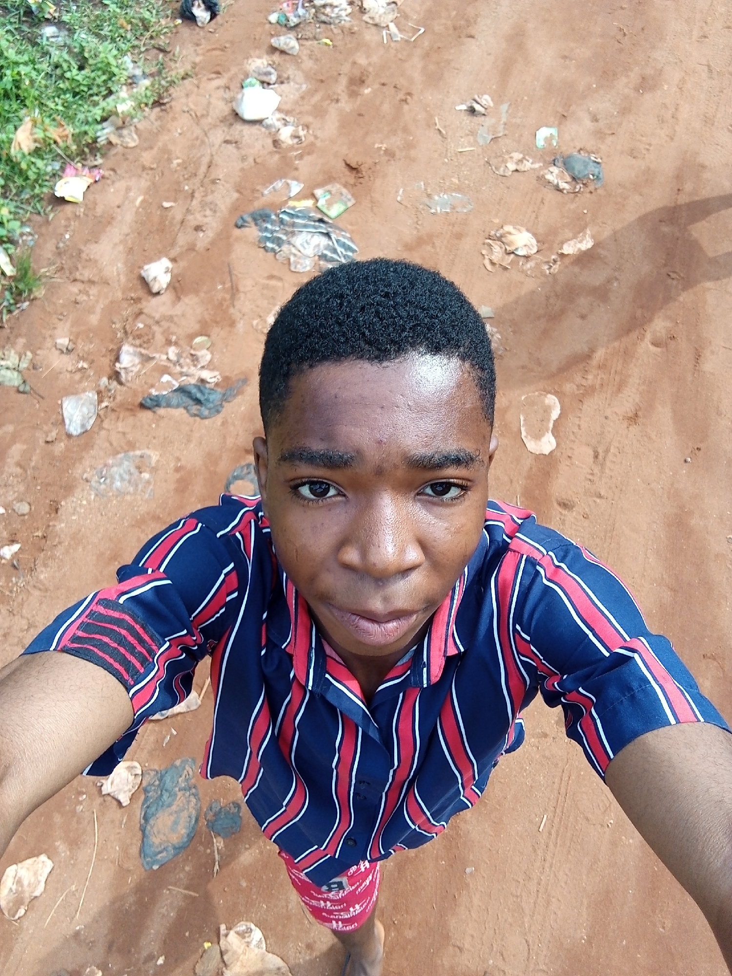 A person, appearing to be a young adult male, is centered in the frame, taking a selfie from a high-angle perspective. The subject has short, dark hair and is wearing a short-sleeved shirt with vertical red and white stripes against a blue background.
The setting is Ukwu-Abwa, Nigeria, as indicated by the sandy soil with visible litter strewn across the ground, including scattered debris, rocks, and possibly discarded packaging. Green vegetation appears in the upper left corner of the image, suggesting a roadside or undeveloped area. The overall impression is a scene of urban disrepair or neglect.