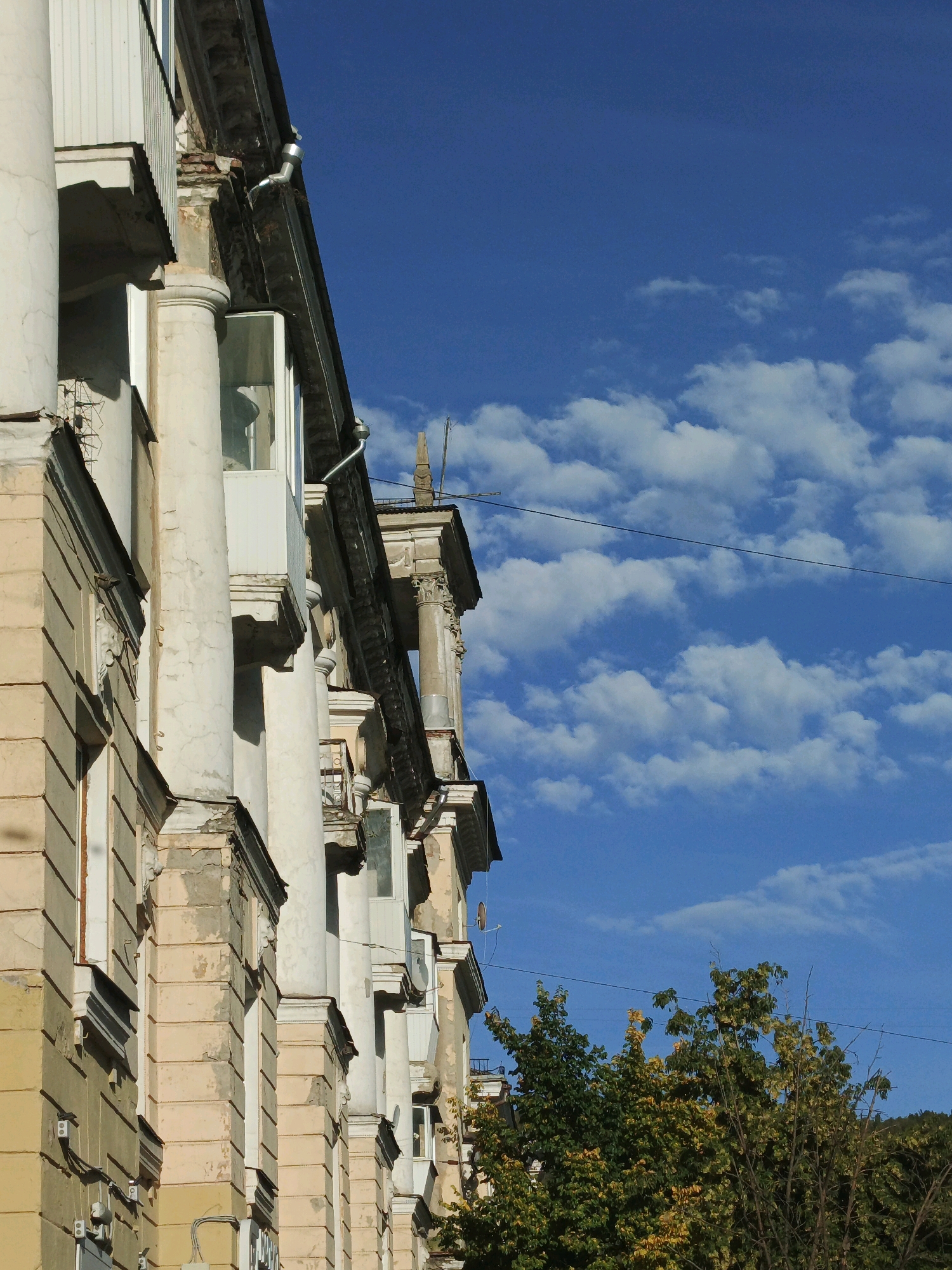 The image captures a close-up view of an old apartment building with classical architectural elements. The building's facade is a pale yellow color, showing signs of age with peeling paint and weathered details. Prominent features include large, fluted columns supporting balconies and ornate cornices. Several windows and balconies are visible, some with glass enclosures. The building appears to be multi-story.
The sky above is a vibrant blue, dotted with fluffy white clouds, suggesting a pleasant daytime weather. A thin black wire stretches across the sky, partially obscuring the building's upper sections and the sky. To the right of the building, a lush green tree with hints of yellow foliage is visible, indicating the possibility of an autumn season. In the far background, a forested hillside is faintly discernible under the clear sky.
The scene evokes a sense of history and the passage of time, with the weathered building standing in contrast to the bright, dynamic sky and foliage. No people are visible in the image. The time of day appears to be late morning or early afternoon, given the lighting and the clear, bright sky. There is no visible text in the image. The location context suggests this is an urban residential building in Zlatoust, Russia.