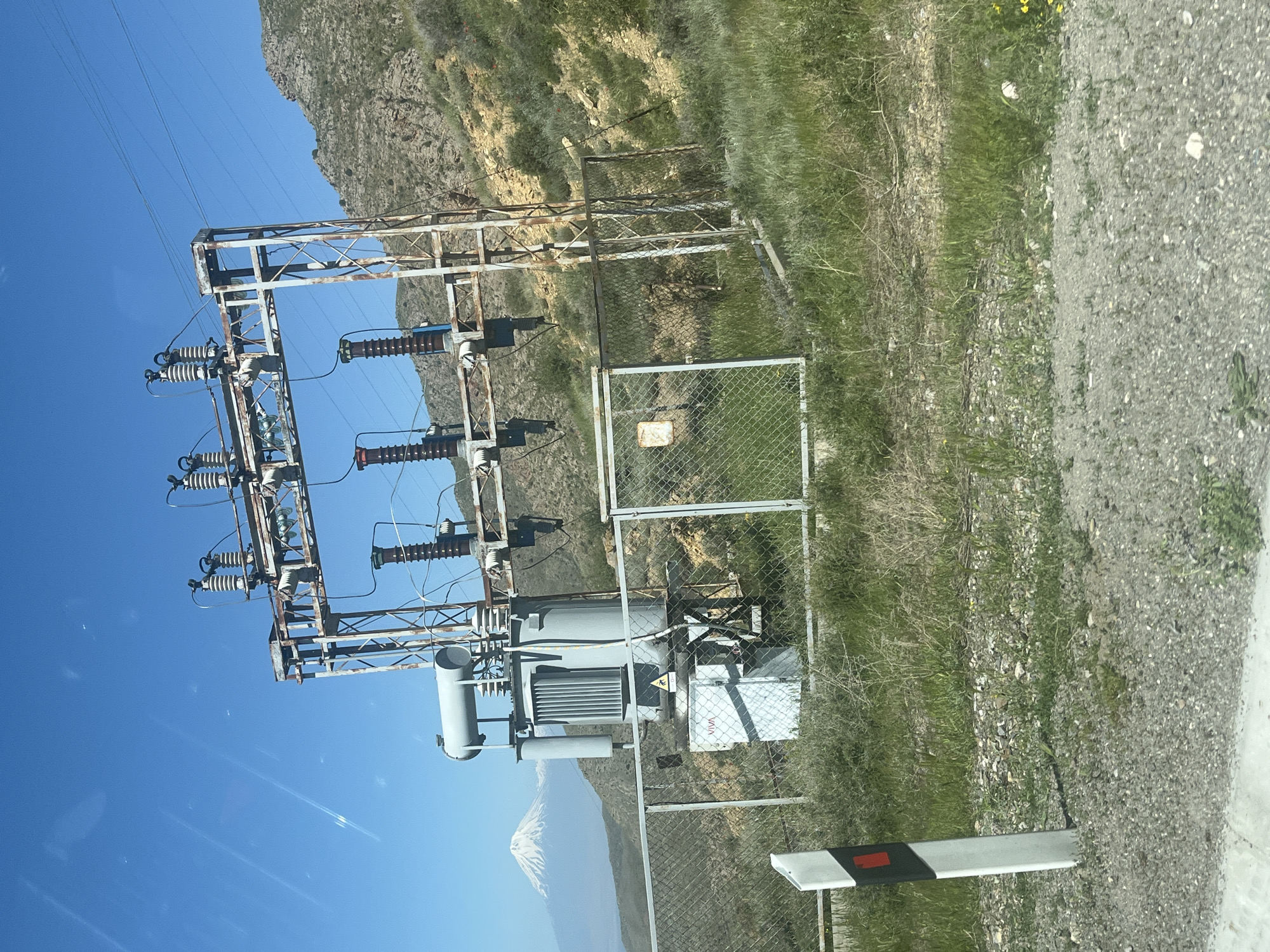 This outdoor photograph, taken on a clear, sunny day, captures an electrical substation set against a backdrop of mountains and a bright blue sky. The image is taken from a low angle, possibly from a vehicle, looking towards the substation and the distant landscape.
In the foreground, a gravel road or shoulder is visible on the right, alongside an area of dry grass and sparse vegetation. A white and black striped marker post is partially visible at the bottom right edge.
The central focus is an electrical substation, featuring a weathered metal framework structure that supports various electrical components, including numerous white ceramic insulators, circuit breakers, and power lines. Many of the metal beams show signs of rust. Below this structure stands a large, gray electrical transformer with cooling fins and a cylindrical conservator tank. The entire substation equipment is enclosed by a chain-link fence, which has a closed gate on the right side. Inside the fenced area, next to the transformer, there's a white electrical cabinet or box with the word "VIVA" clearly visible on it, along with a yellow triangular warning sign indicating danger.
Behind the substation, rocky hills with patches of dry grass and sparse vegetation extend across the mid-ground. In the far distance, prominent against the clear blue sky, is a majestic snow-capped mountain. Its upper slopes are covered in white snow, while the lower parts appear dark, contrasting sharply with the sky.
The sky is a brilliant, cloudless blue, indicating clear weather. The bright light suggests it is daytime, likely mid-morning or afternoon. There are no people visible, and no specific activity is taking place. The scene presents a blend of industrial infrastructure and natural landscape.