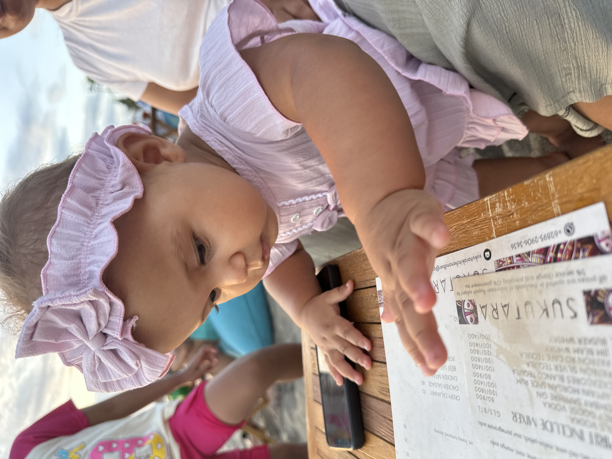 In this close-up, slightly tilted image, a baby wearing a pink ruffled headband and a pink dress is the primary subject. The baby's face is turned towards the right side of the frame, with eyes looking down at a menu on a wooden table. The baby's left hand is reaching out towards the menu. To the left of the baby, an adult wearing a white t-shirt is partially visible, and to the right, another person is partially visible, wearing a light gray garment. The setting appears to be outdoors, possibly a restaurant or beachside cafe, given the wooden table and the natural light. The sky is visible in the background.
The menu on the table contains various food and drink items, with prices listed in what appears to be Indonesian Rupiah, as indicated by the text "+62895-2906-3636" and "Indonesian Rupiah". Some of the visible text on the menu includes "SUKUTARA", "MIXER", "VODKA", "GIN", "TEQUILA", "RUM", "WHISKY", "CHICKEN SAUSAGE", and "CRISPY CALAMARY". The time of day seems to be late afternoon or early evening, judging by the warm, soft lighting. The overall mood of the image is candid and intimate, focusing on the baby's curiosity and interaction with the menu.