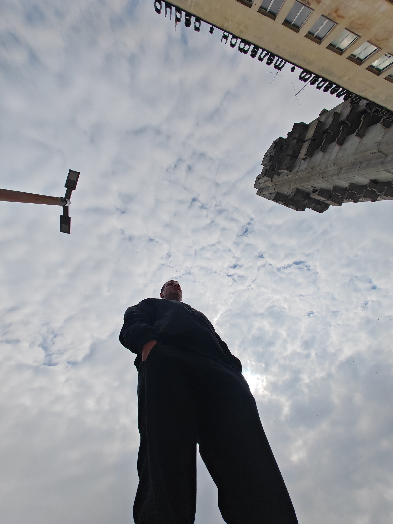The image displays a low-angle perspective of an urban environment in Shumen, Bulgaria, under an overcast sky with scattered clouds. A single adult male, partially silhouetted against the bright sky, stands in the lower central frame. He wears dark, long-sleeved attire and trousers, with hands appearing to be in his pockets, and is observed looking upwards towards the upper right. To the upper left, a dark, T-shaped street light fixture with two rectangular luminaires extends horizontally. In the upper right quadrant, two architectural structures are visible: a light-colored building with multiple rectangular windows displays inverted dark Cyrillic letters spelling "МАРШАН" (MARSHAN) on its lower facade. Adjacent to and partially below this building, a large, grey concrete structure featuring a brutalist style with prominent geometric, block-like protrusions is visible, extending from the upper right corner.