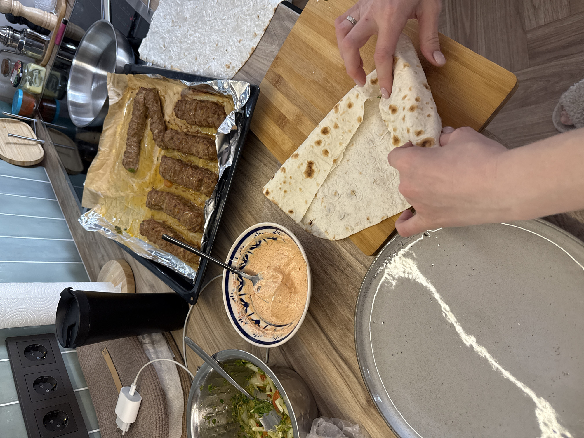 The image shows a meal preparation scene, possibly taking place in a kitchen or dining area. A person is in the process of folding a flatbread, likely a lavash or similar type. Nearby, there is a baking sheet with cooked meat kebabs, a bowl with what appears to be a dipping sauce, and another bowl containing a salad. A plate is visible, presumably for serving the prepared food. A cutting board sits next to the person folding the flatbread. Other items present include kitchenware, a charger, and a tumbler. The location is assumed to be Krasnoyarsk, Russia.
