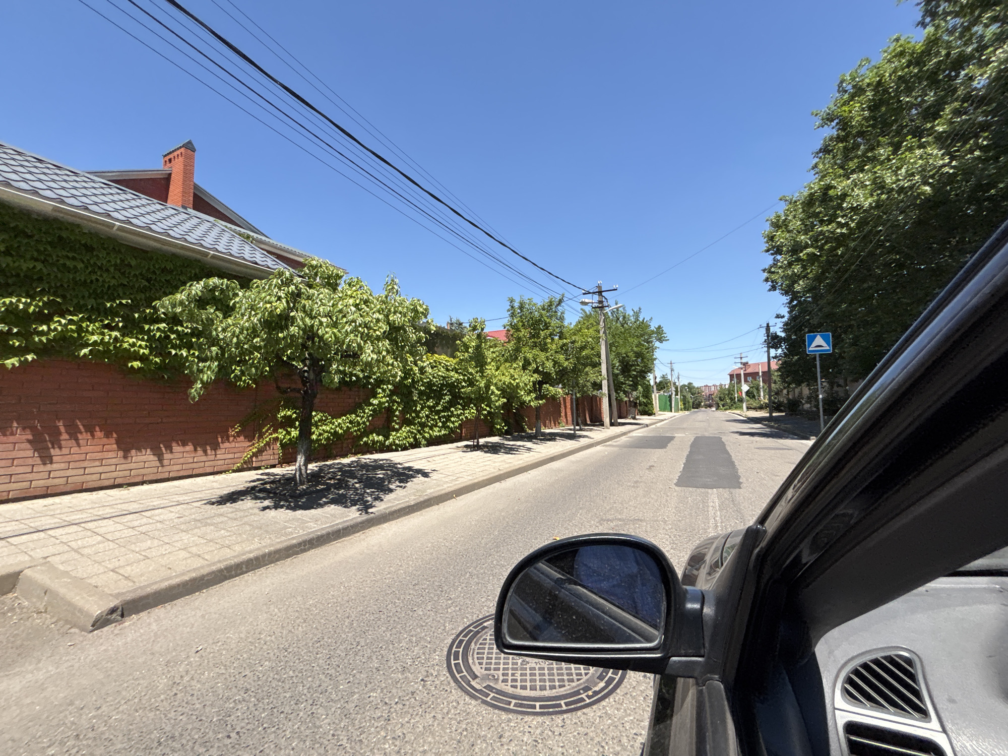The image captures a view from the passenger side of a vehicle traveling down a paved street in a residential area, likely in Krasnodar, Russia, under a clear blue sky. The street is lined with sidewalk and a brick wall covered in ivy on the left, with a residential building with a red brick chimney and a grey tiled roof visible behind it. Green trees with dense foliage are prominent along the sidewalk, casting shadows on the pavement. A traffic sign with a blue triangle and a white symbol is visible on the right side of the street. The vehicle's side mirror is in the foreground, reflecting the sky and a portion of the car's interior. A manhole cover is visible on the road near the mirror. The road surface shows some patched areas. The overall impression is of a sunny day in a suburban or residential part of a city.