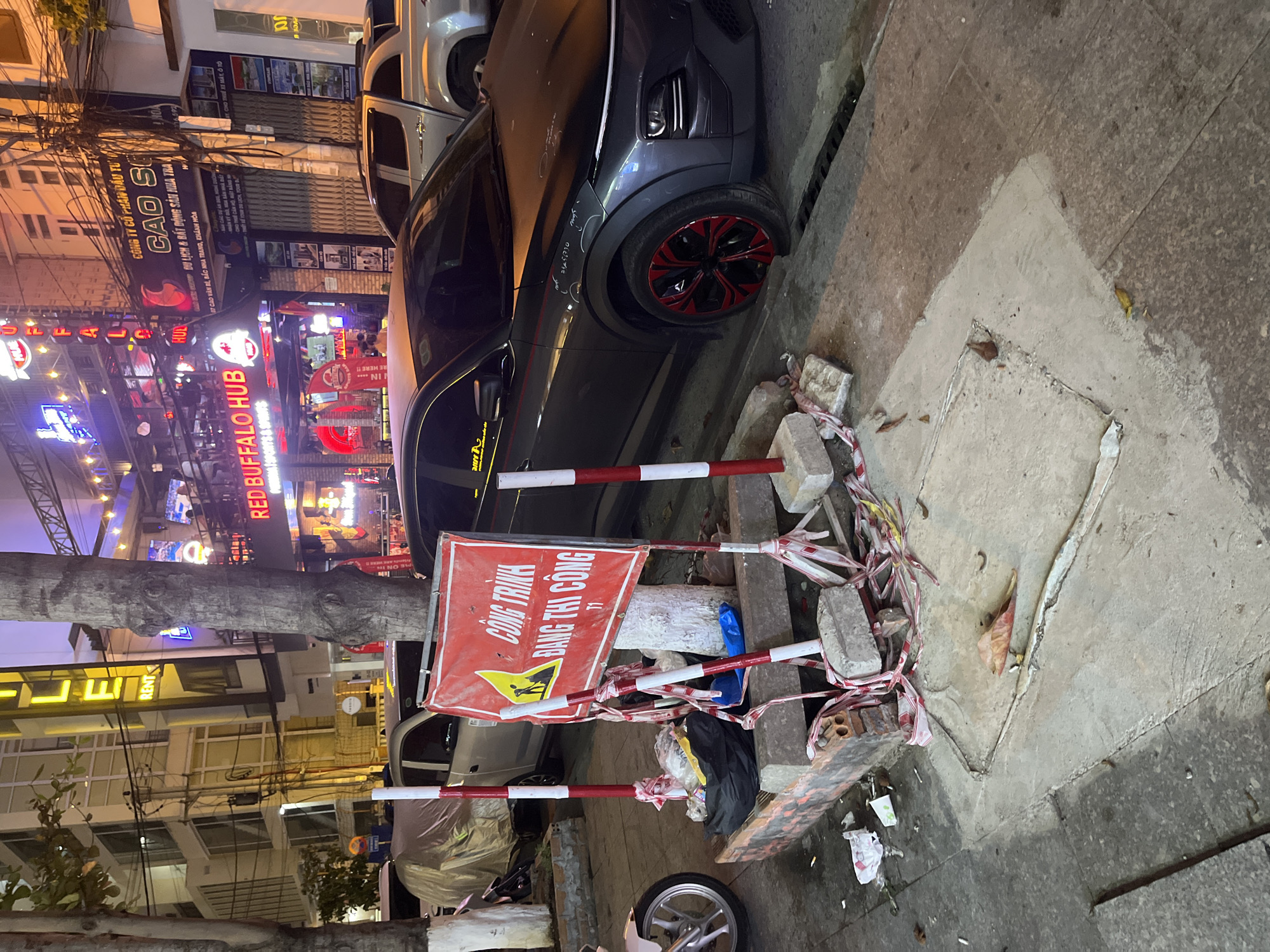 The image depicts a street scene in Bac Nha Trang Ward, Vietnam, at night. A dark grey car with red-accented wheels is partially parked on the sidewalk, encroaching on a construction area marked by a red and white barrier. The barrier is surrounded by bricks, debris, and caution tape. A sign in Vietnamese reads "CÔNG TRÌNH ĐANG THI CÔNG," indicating ongoing construction. In the background, illuminated signs for "RED BUFFALO HUB" and other businesses are visible. A tree trunk is positioned near the construction area. Other parked vehicles and buildings line the street. The sidewalk is concrete with visible repairs.