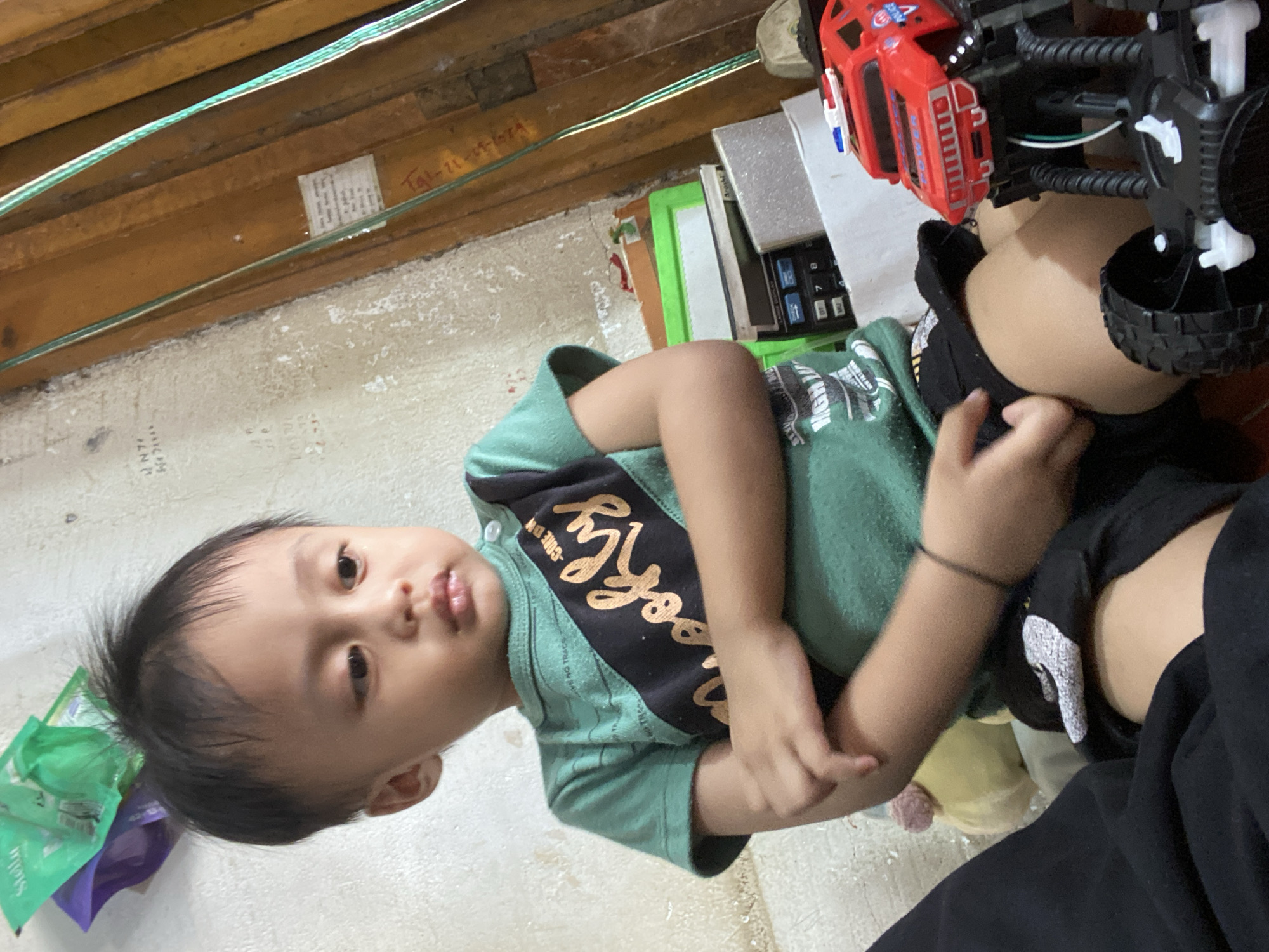 In the city of Ta', Indonesia, a young boy is seated on the floor, looking down and to his right with his arms crossed. He is wearing a green t-shirt with a black diagonal stripe that says "Brooklyn" in gold lettering, and black shorts. In front of him, to his right, is a red toy police truck. The background consists of a textured, light-colored wall and a wooden structure with wires running down it. There are also several stacked items behind the boy, including what appears to be a calculator and boxes. A yellow plush toy is visible to the boy's left.