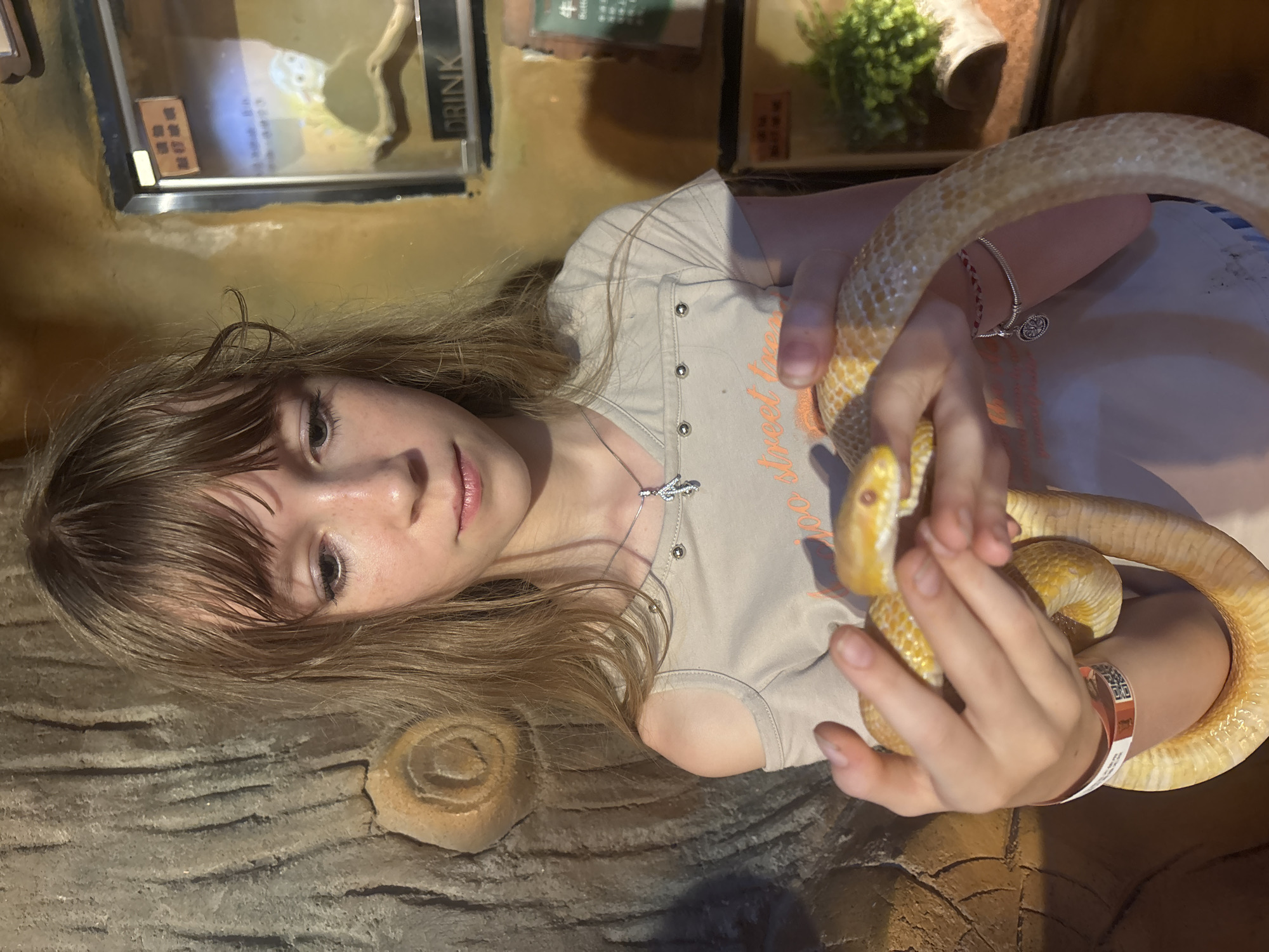 A young girl with blonde hair and bangs is holding a yellow corn snake in her hands. She is wearing a light-colored t-shirt with an off-the-shoulder design and a necklace with a pendant. The snake is wrapped around her forearms and is yellow with orange markings. The background appears to be a zoo or aquarium exhibit, with a textured, rocky wall on the left and glass enclosures with plants and signage on the right. The signage on the right includes Chinese characters and the word "DRINK". The girl is looking down at the snake with a neutral expression. A wristband with a QR code is visible on her left wrist. The overall lighting suggests an indoor environment, possibly with artificial lighting. The location is the zoo in Tianhe District, China.