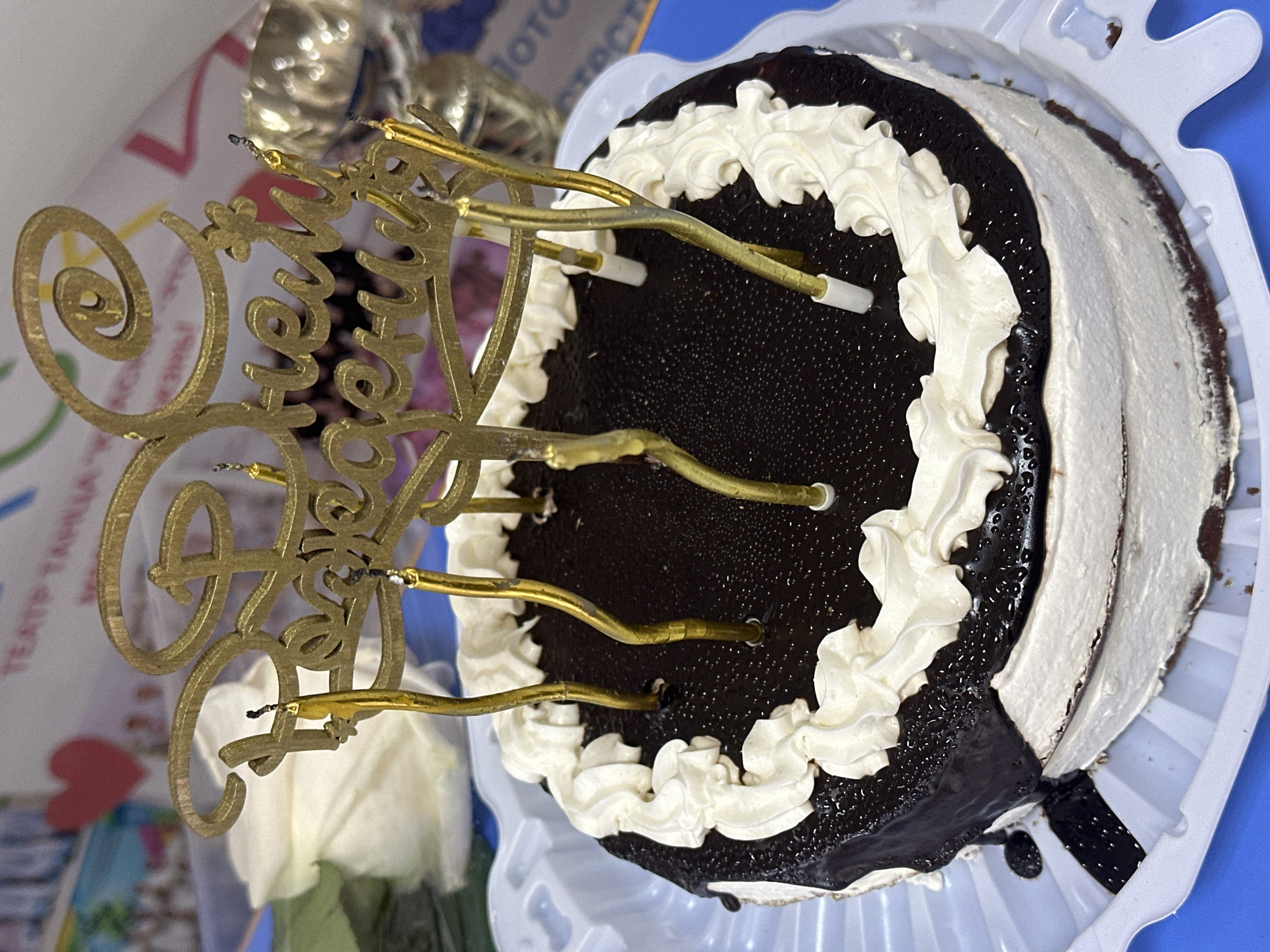 A chocolate cake decorated with white frosting and a golden "Happy Birthday" topper sits on a white plastic tray, likely in Votkinsk, Russia, indicated by lettering on a partial background item. The cake is two-toned, split roughly in half with chocolate ganache on one side and white frosting on the other, separated by a swirl of white whipped cream on top. Six golden candle holders stick out of the cake. The background reveals partial text and a gold-colored object and a white flower. The cake is set on a blue surface.