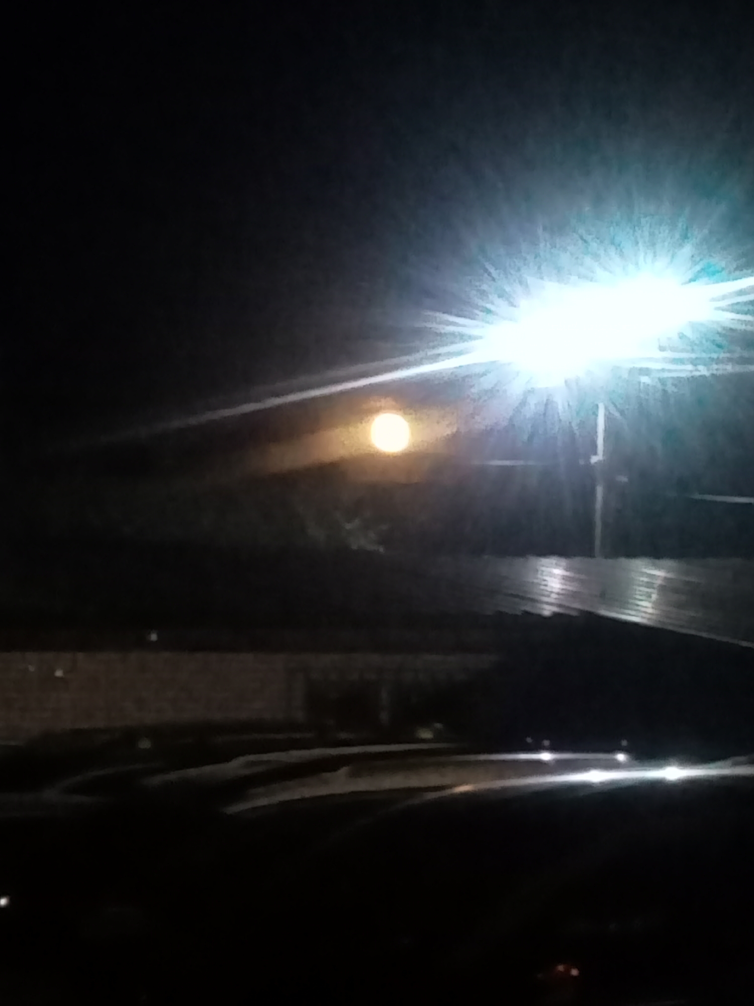 A nighttime image captures a bright overhead light source with visible rays, positioned above and to the right of a full moon. The moon, illuminated and slightly hazy, is located in the upper central portion of the frame, casting a faint orange glow. Below these celestial and artificial lights, a dark, ribbed surface, possibly a roof, spans the mid-ground. In the foreground, a dark, indistinct area with scattered horizontal white light streaks suggests the presence of vehicles or a roadway at night. The background is predominantly dark, with some subtle, shadowy outlines of trees or structures behind the moon. The overall scene depicts an urban or semi-urban environment at night, illuminated by both natural and artificial light.